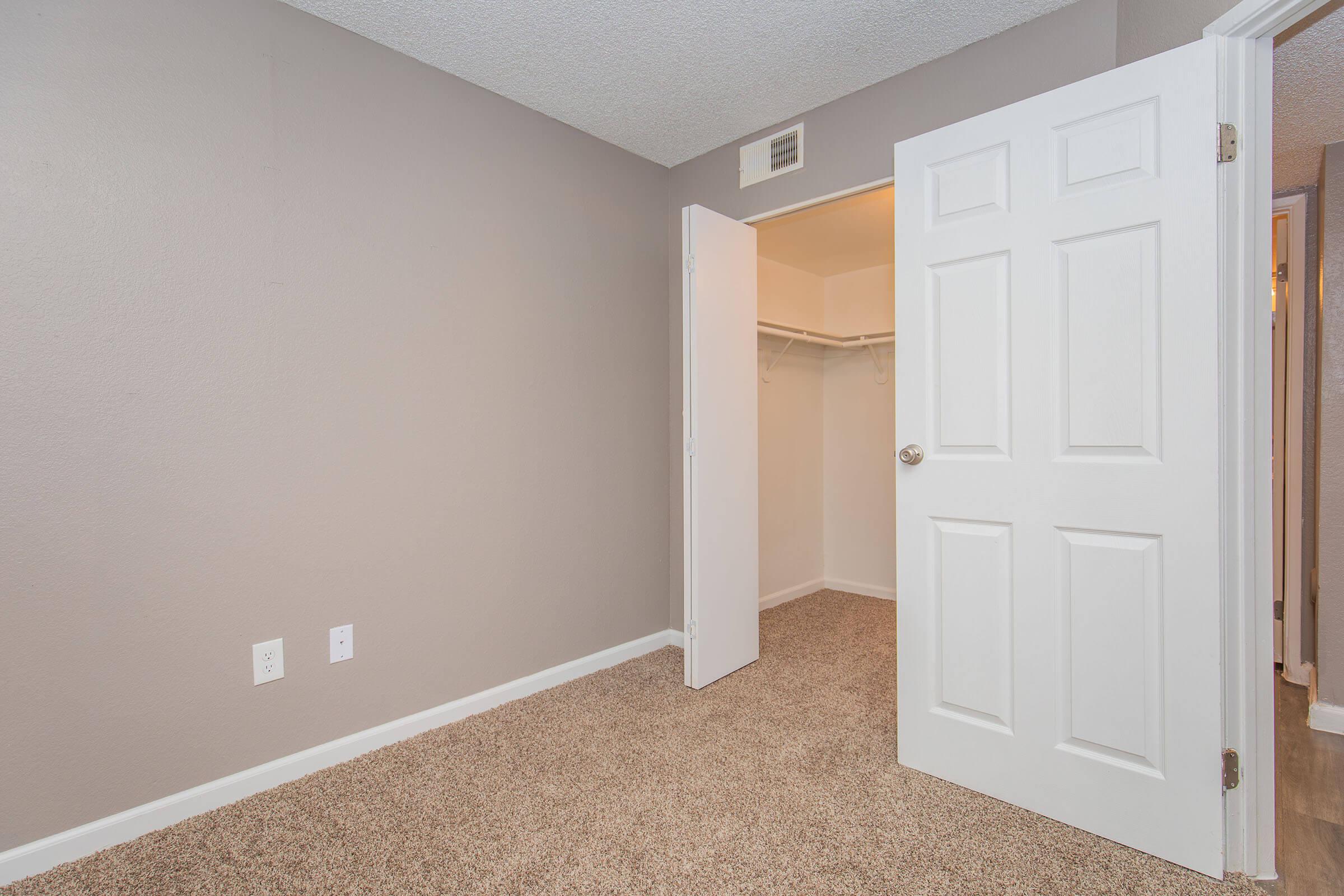 Empty room with light gray walls and beige carpet. A white door is slightly open, revealing a small closet with no visible contents. Two electrical outlets are visible on the wall. The overall appearance is clean and well-lit, suggesting a recently updated space.
