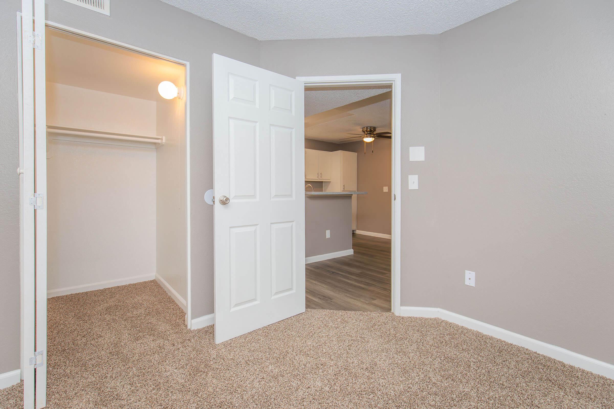 Interior view of a room featuring a beige carpet, a closet to the left with an open door, and a doorway leading to an adjacent space. The walls are painted in a light gray tone, and there's a ceiling fan visible in the background. Natural light enters from a nearby window.