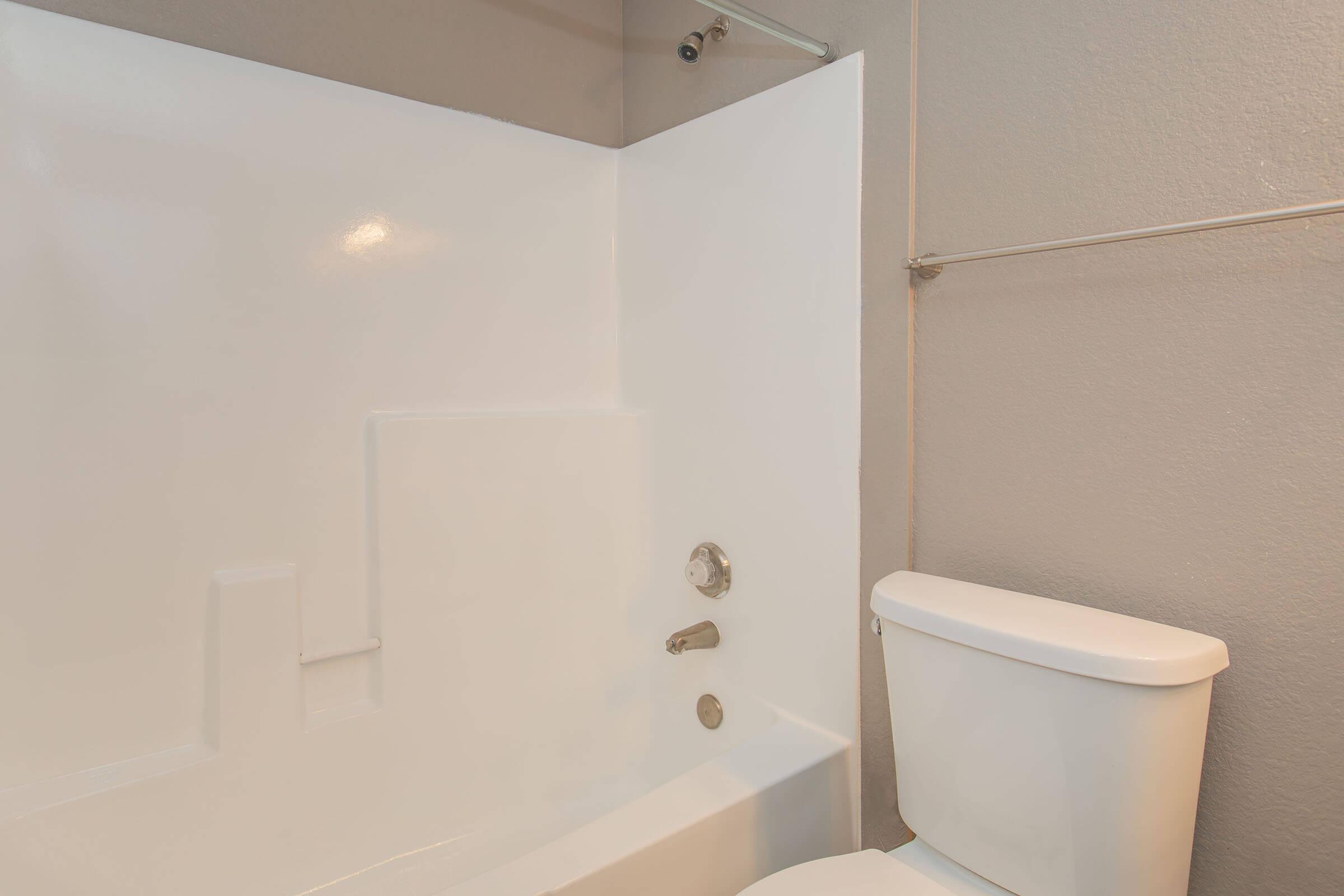 A clean, modern bathroom featuring a white bathtub and shower combo, with a chrome faucet and wall-mounted showerhead. Adjacent is a white toilet. The walls are painted in a neutral tone, providing a bright and open feel to the space.