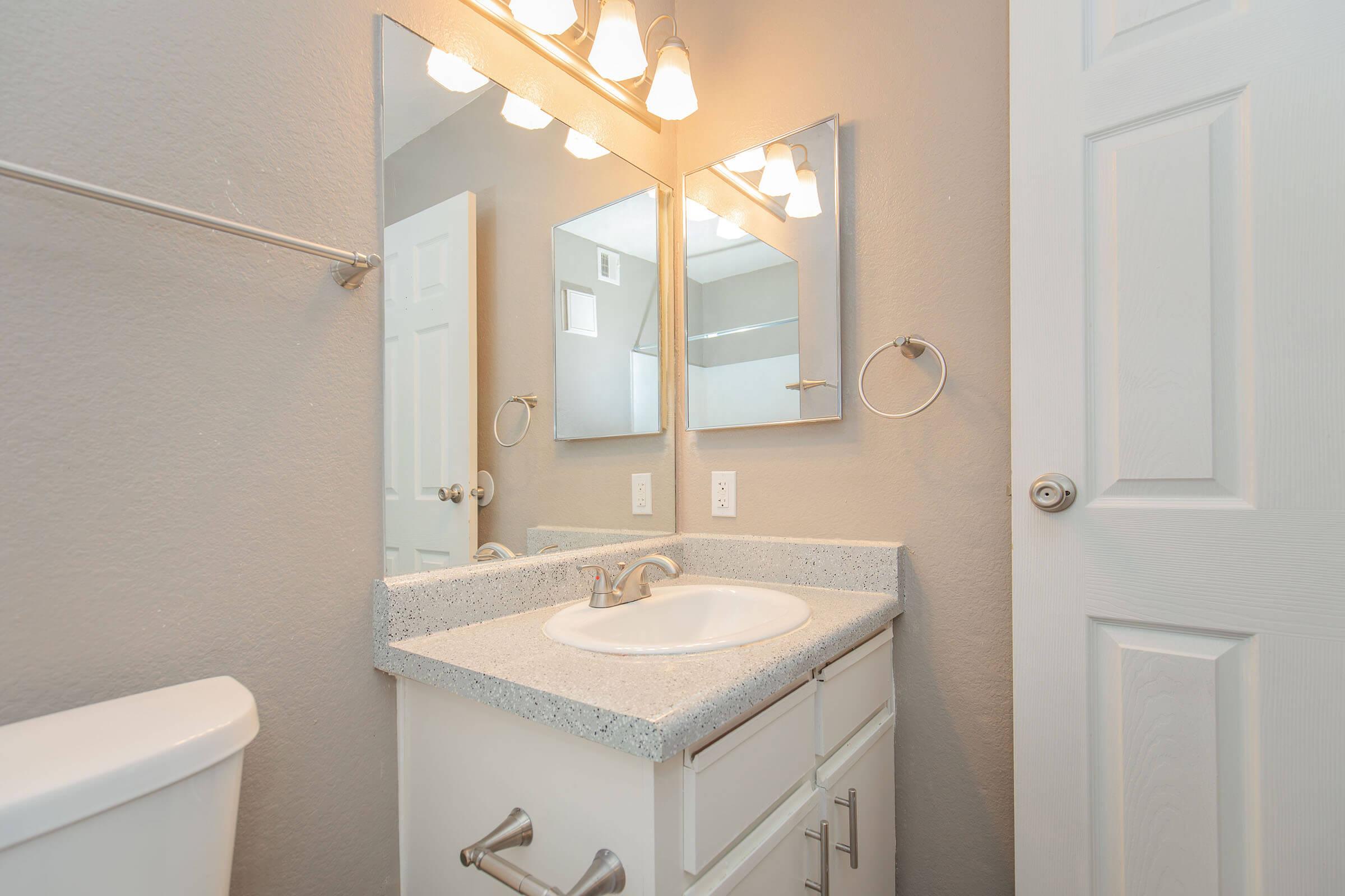 A modern bathroom featuring a white countertop with a sink, two mirrors above it, a towel holder on the wall, and a light fixture with three bulbs. The walls are painted gray, and a white door is visible in the background next to a toilet. Bright lighting enhances the clean and contemporary look of the space.