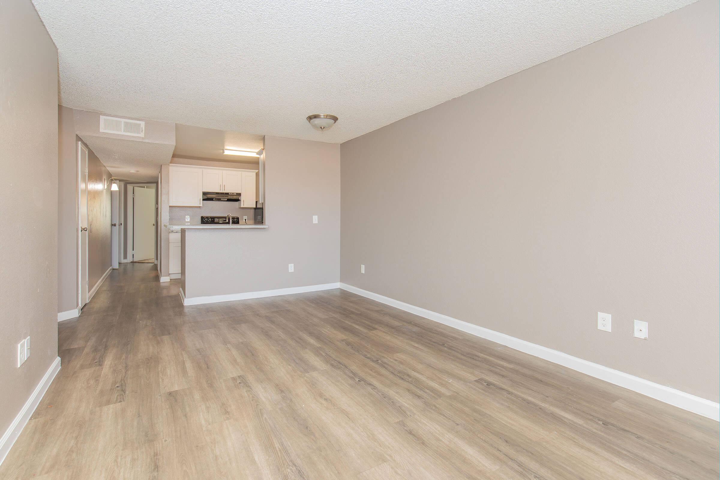 Spacious multi-purpose room with light-colored walls and laminate flooring. The area is empty, featuring a ceiling light fixture. In the background, a kitchen with white cabinetry and a hallway is partially visible, leading to an entrance. Natural light brightens the space, creating an inviting atmosphere.