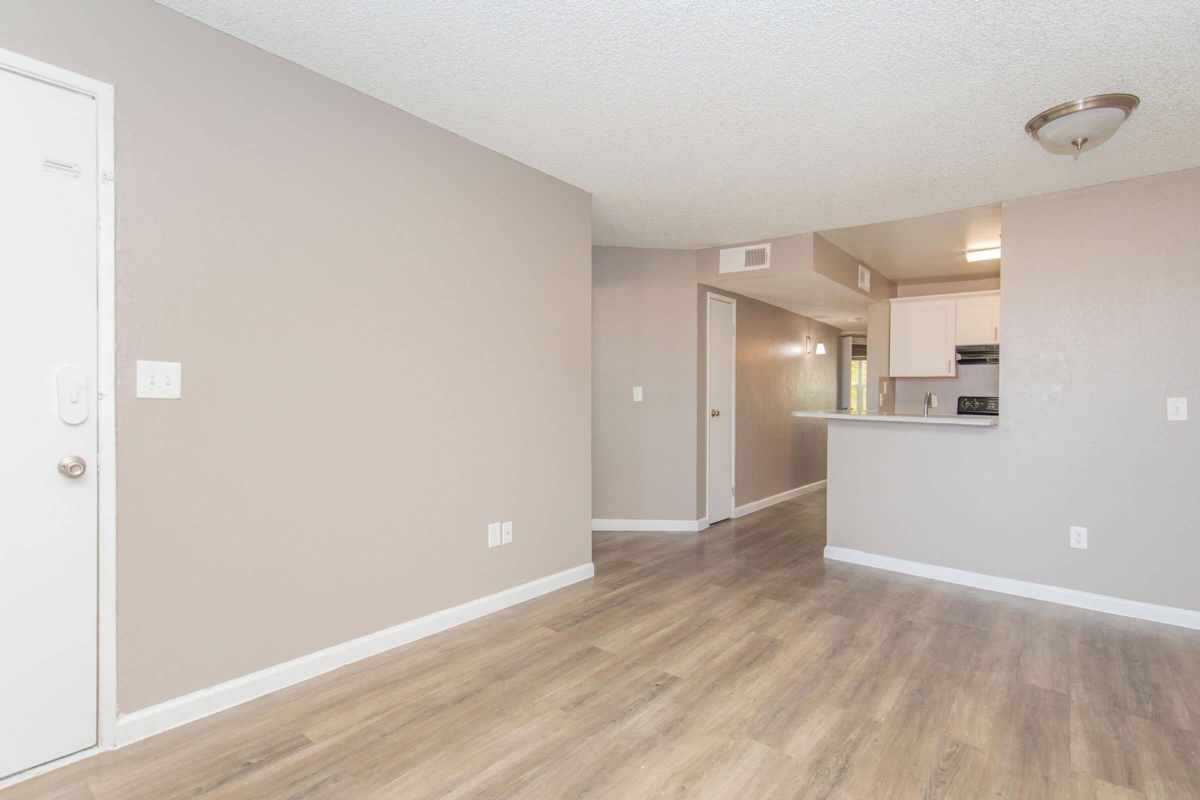 Interior view of a modern apartment featuring a spacious living area with light gray walls and wood-like flooring. A doorway on the left leads to the entrance, while an open kitchen with white cabinets and appliances is visible in the background. Natural light emanates from nearby windows.