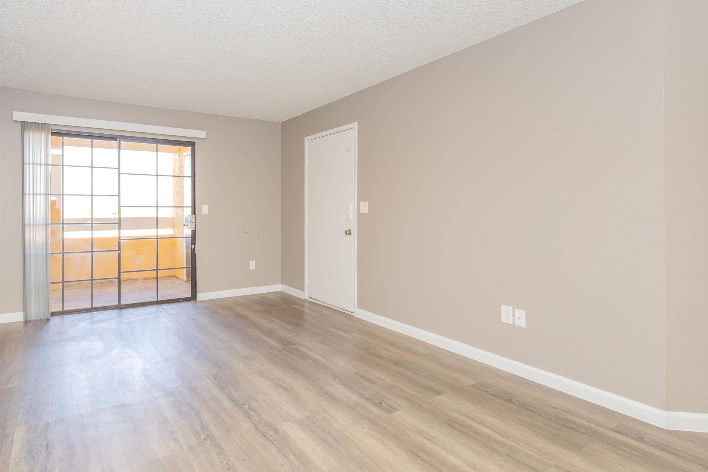 An empty room with light-colored walls and wooden flooring. A glass sliding door leads to a balcony, letting in natural light. There is a white door on the right side of the image. The overall atmosphere is bright and spacious, suitable for various interior designs.