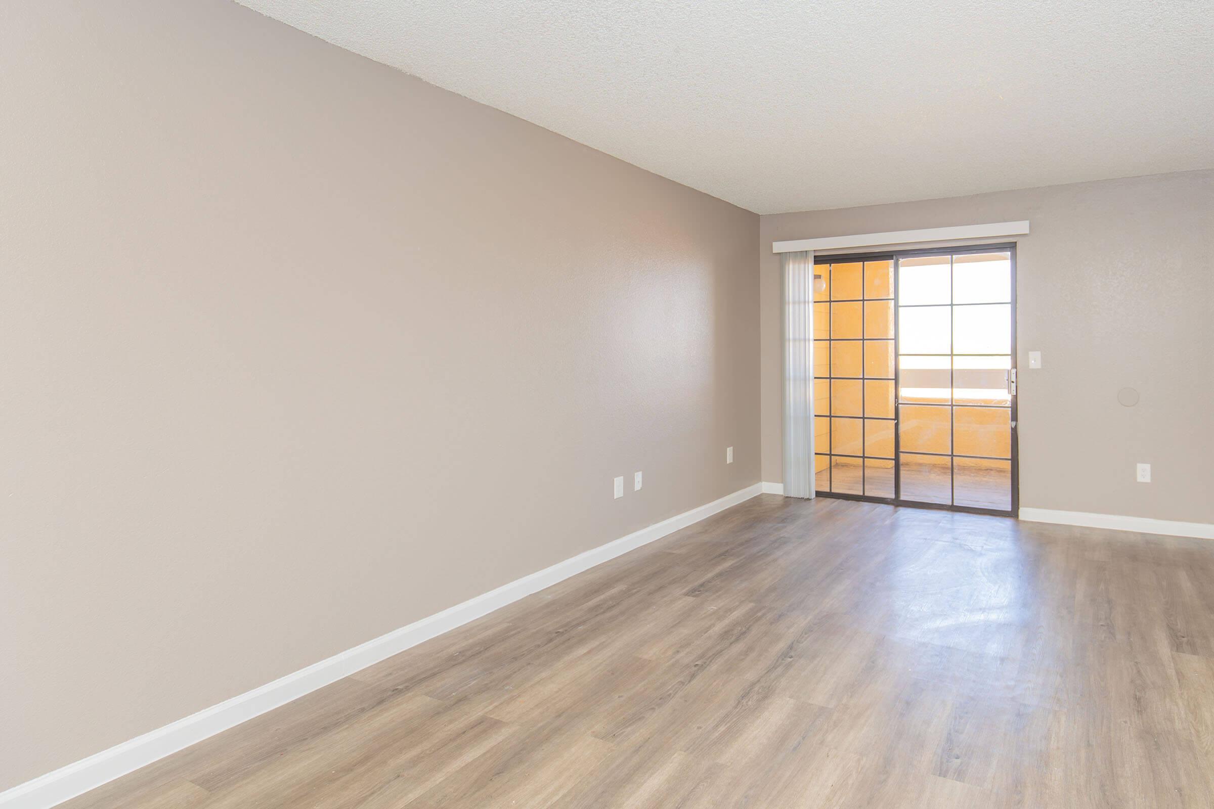 A spacious, empty room with light beige walls and wooden flooring. A sliding glass door provides access to a balcony, allowing natural light to enter the space. The overall atmosphere is bright and open, suitable for various interior design possibilities.
