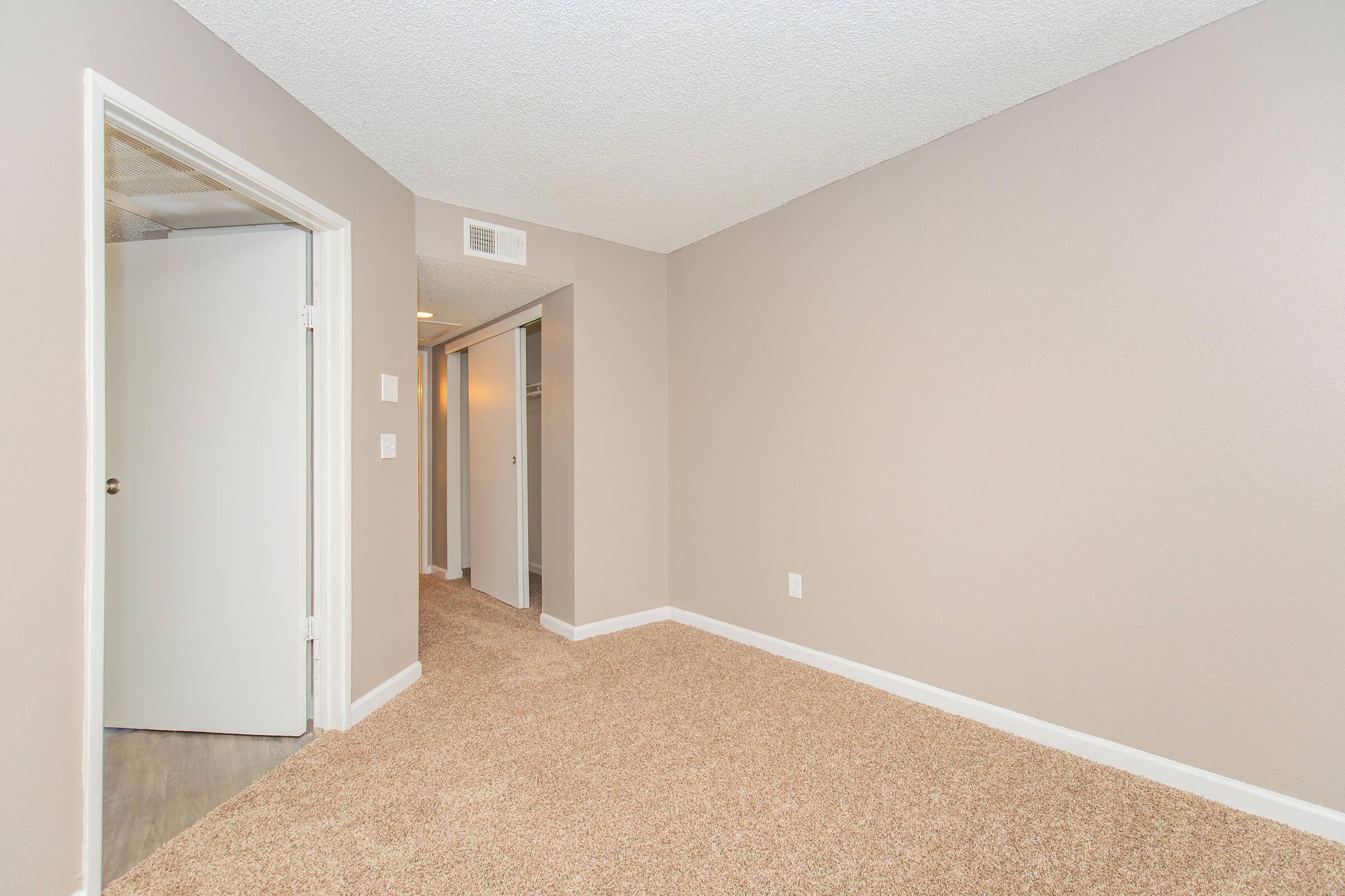 A light beige carpeted room with neutral-colored walls. A doorway on the left leads to another room, while a hallway is visible at the far end. The ceiling has a textured finish, and a small closet is partially visible in the hallway. Natural light enters the space, creating a bright atmosphere.