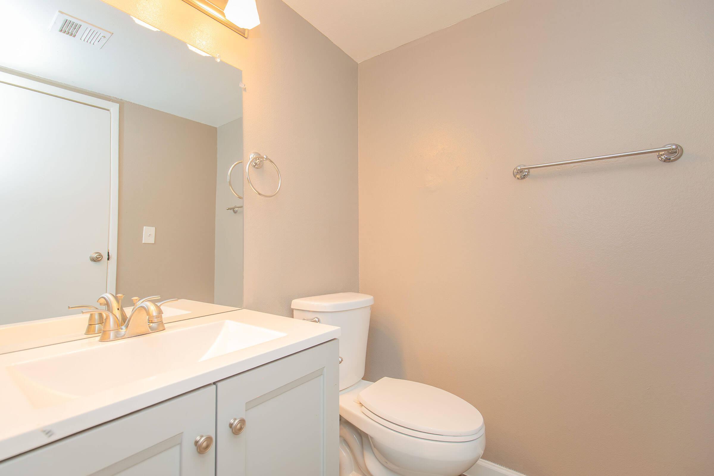 A clean bathroom with a light gray wall. It features a white vanity with a sink and modern faucet, a white toilet, and a wall mirror. A towel bar is mounted next to the sink, and there is a door leading to another space in the background. The overall ambiance is bright and minimalistic.