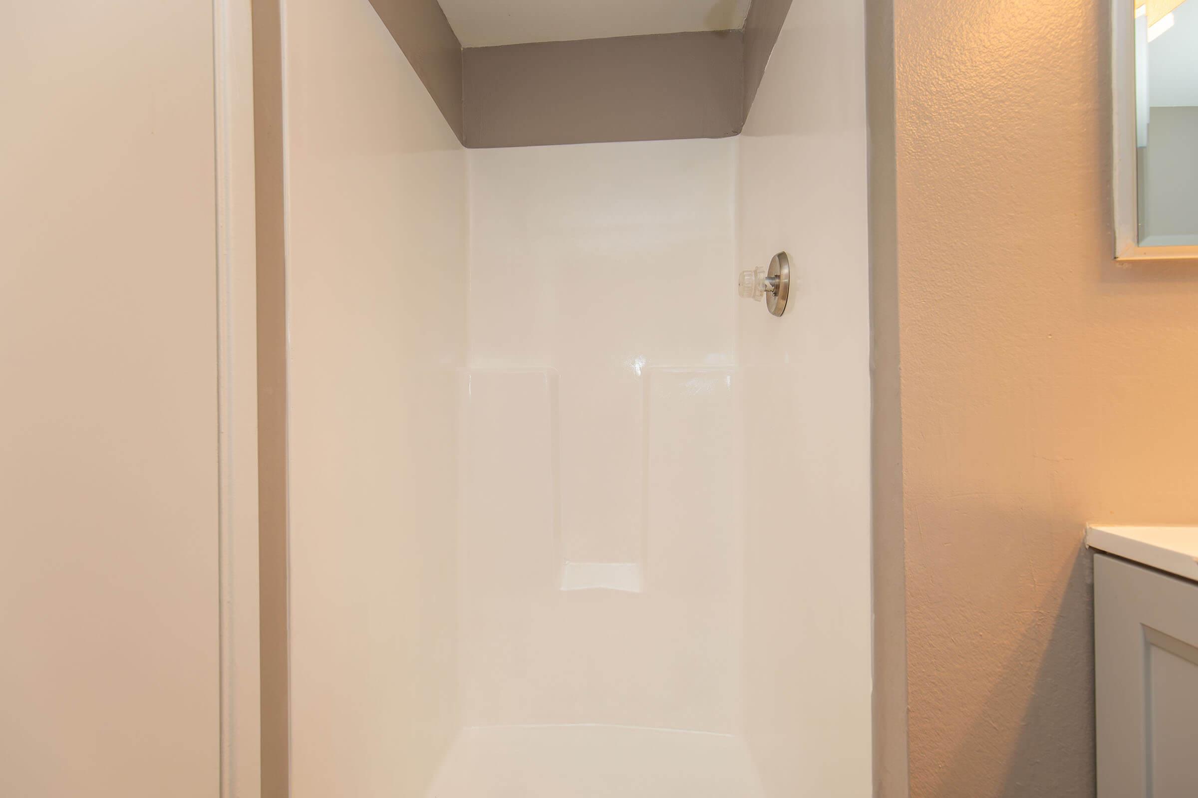 A clean, modern shower stall featuring a smooth white interior and a single chrome faucet. The shower is surrounded by neutral-colored walls, with a partly visible vanity in the background. Soft lighting enhances the bright and spacious feel of the bathroom area.