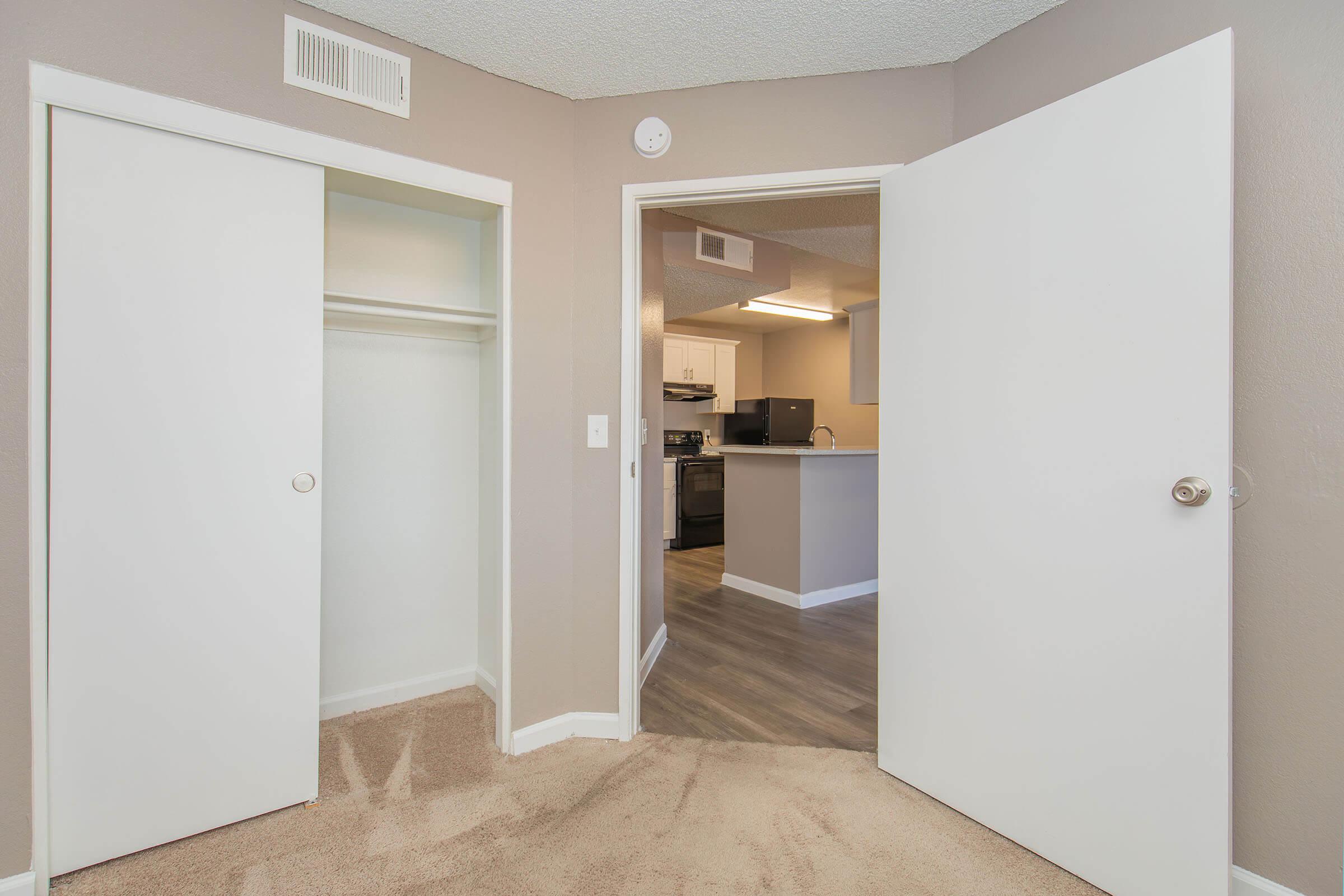 Interior view of a room showcasing two white sliding closet doors on the left and an open door leading to a modern kitchen area with light-colored cabinetry and appliances. The walls are painted in a neutral tone, and the floor has wood-like laminate. The carpeted area adds warmth to the space.