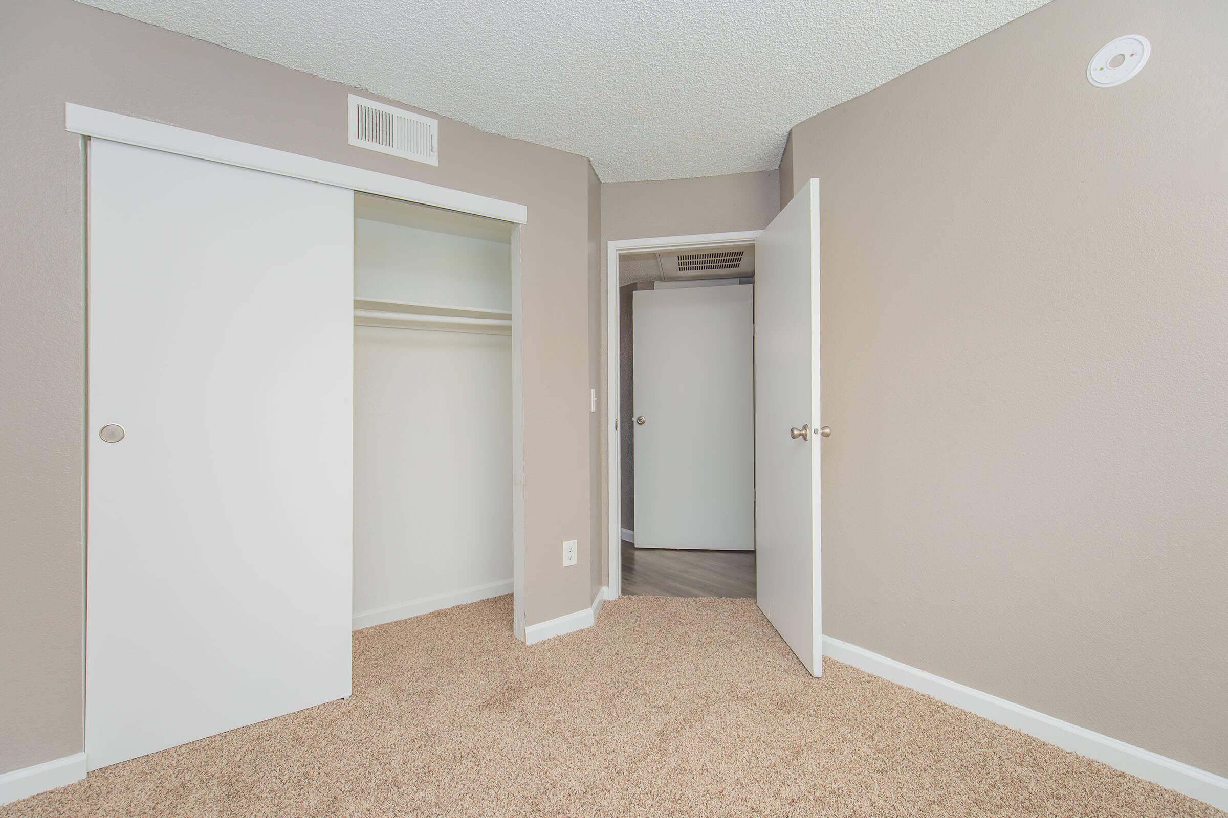 A small room with light brown walls and carpeted flooring. There are two white sliding closet doors on the left and a partially open door on the right leading to another space. The room has a simple, clean appearance, with no furniture visible.