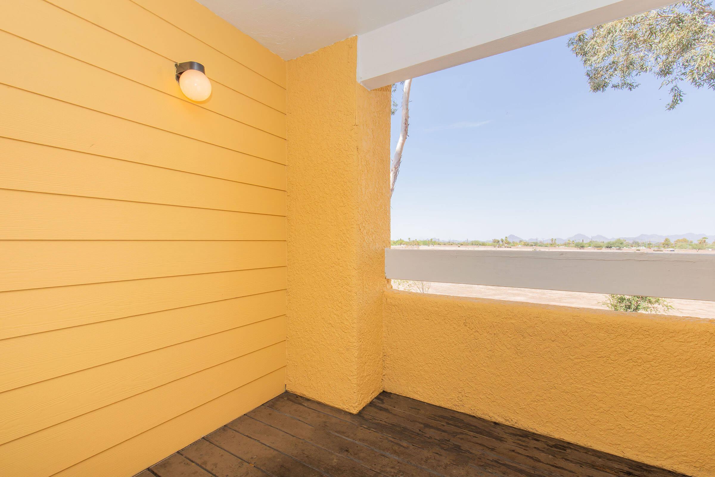 A corner balcony with yellow walls, featuring a light fixture, and an open view of a landscape with distant mountains and a clear blue sky. The floor is made of wooden planks, creating a warm and inviting atmosphere.