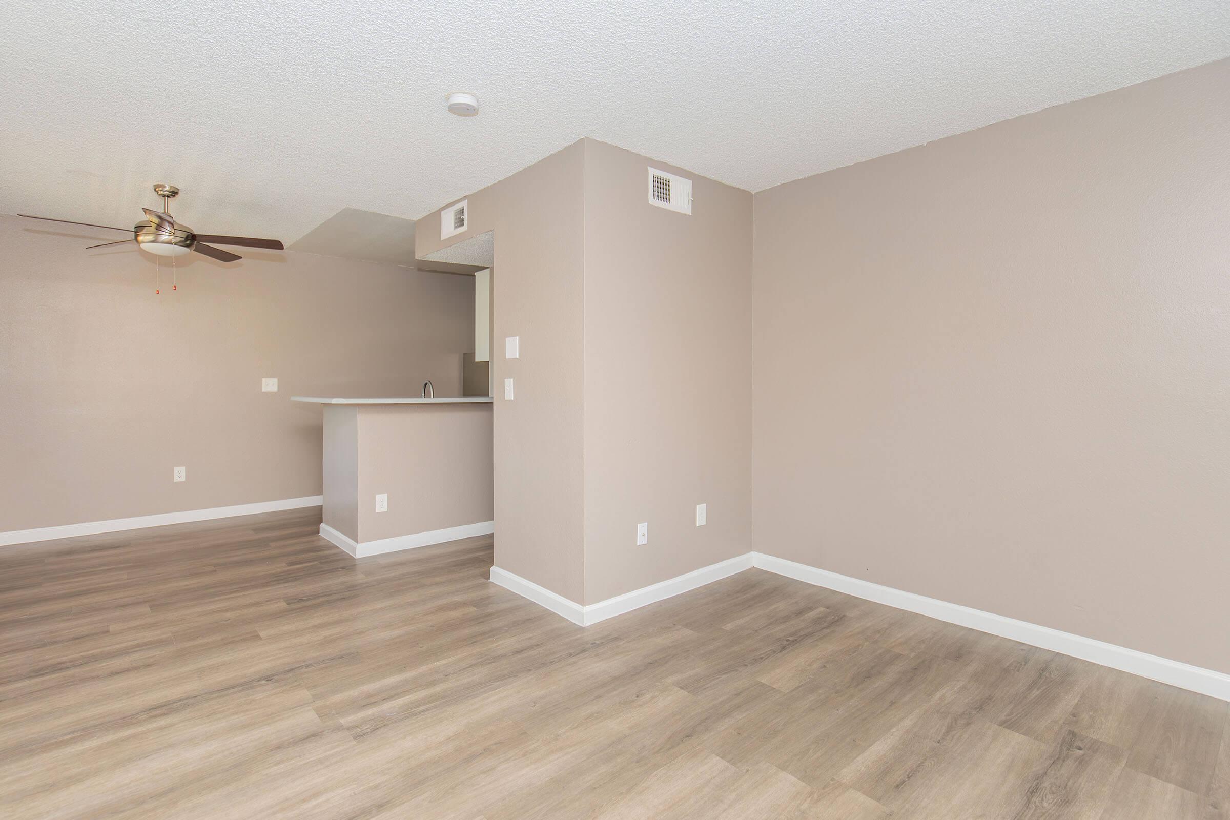 A bright, spacious interior of an apartment featuring light beige walls, a ceiling fan, and wooden flooring. There is a small kitchen area visible in the background with a counter, and empty space suitable for furniture arrangement, creating an open and modern living environment.