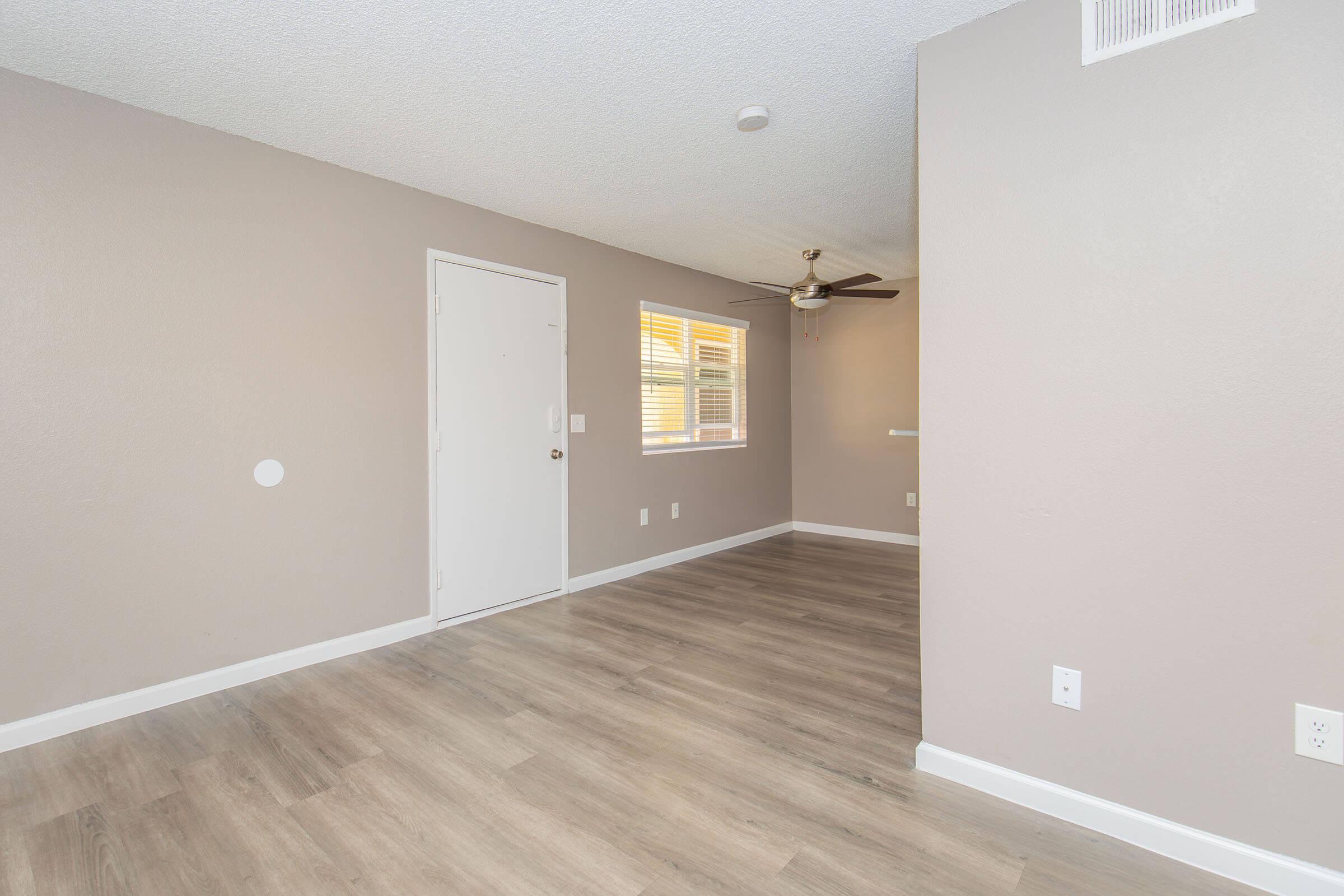 Spacious, well-lit room with light brown walls and wood-style flooring. A front door is visible on the left, and a window with blinds allows natural light in. A ceiling fan hangs from the ceiling, and a shelf is mounted on the wall, creating an inviting atmosphere for living or dining.