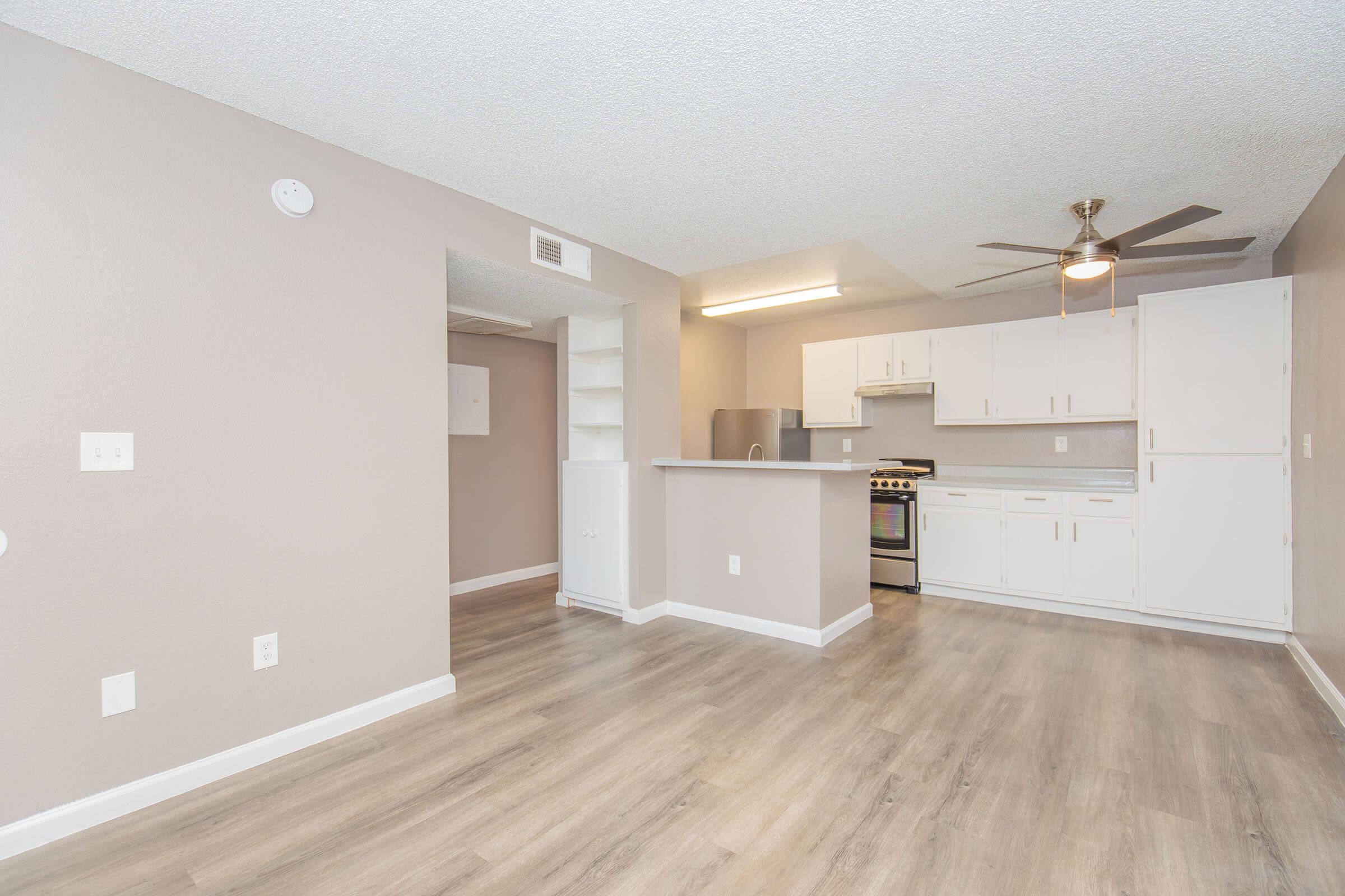 A modern, open-concept living space featuring light-colored walls, a ceiling fan, and laminate flooring. The kitchen area includes white cabinetry and stainless steel appliances. There is a breakfast bar and a small nook with shelves, creating a functional and inviting environment. Natural light brightens the room.