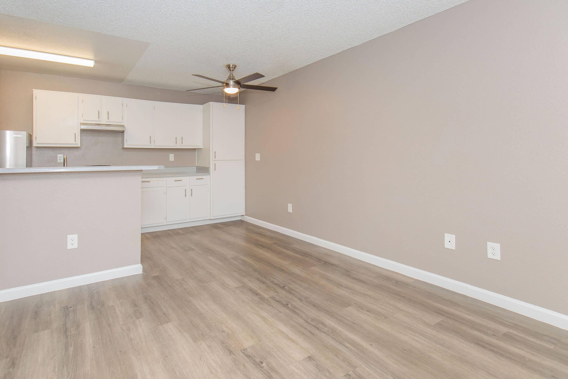 A spacious, well-lit living area featuring light wood flooring, a ceiling fan, and a neutral painted wall. In the background, there's a modern kitchen with white cabinets and a countertop, providing an open-concept layout. No furniture is present in this area.