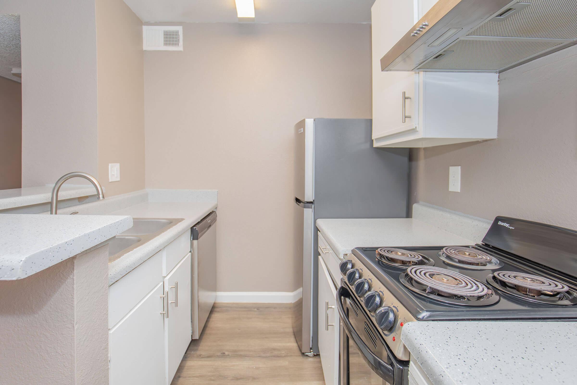 Modern kitchen featuring a stainless steel refrigerator, a sleek stove with four burners, and a double sink. The walls are painted in a soft beige, and the countertops are light-colored with a subtle texture. Hardwood flooring complements the clean and contemporary design of the space.
