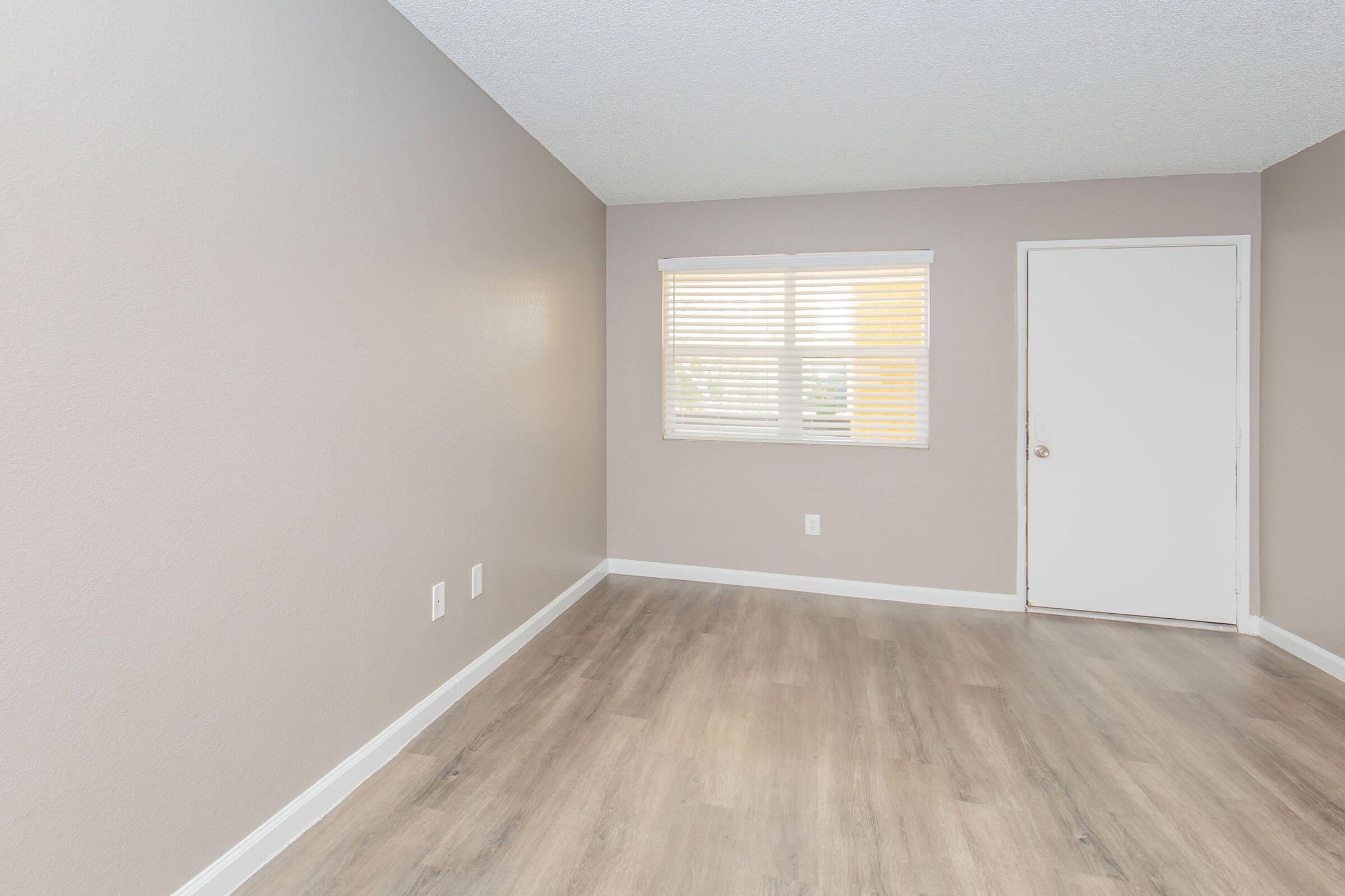 A clean, empty room with light purple walls, large window covered with white blinds, and a door on the right. The floor features light-colored laminate, creating a modern and spacious appearance.