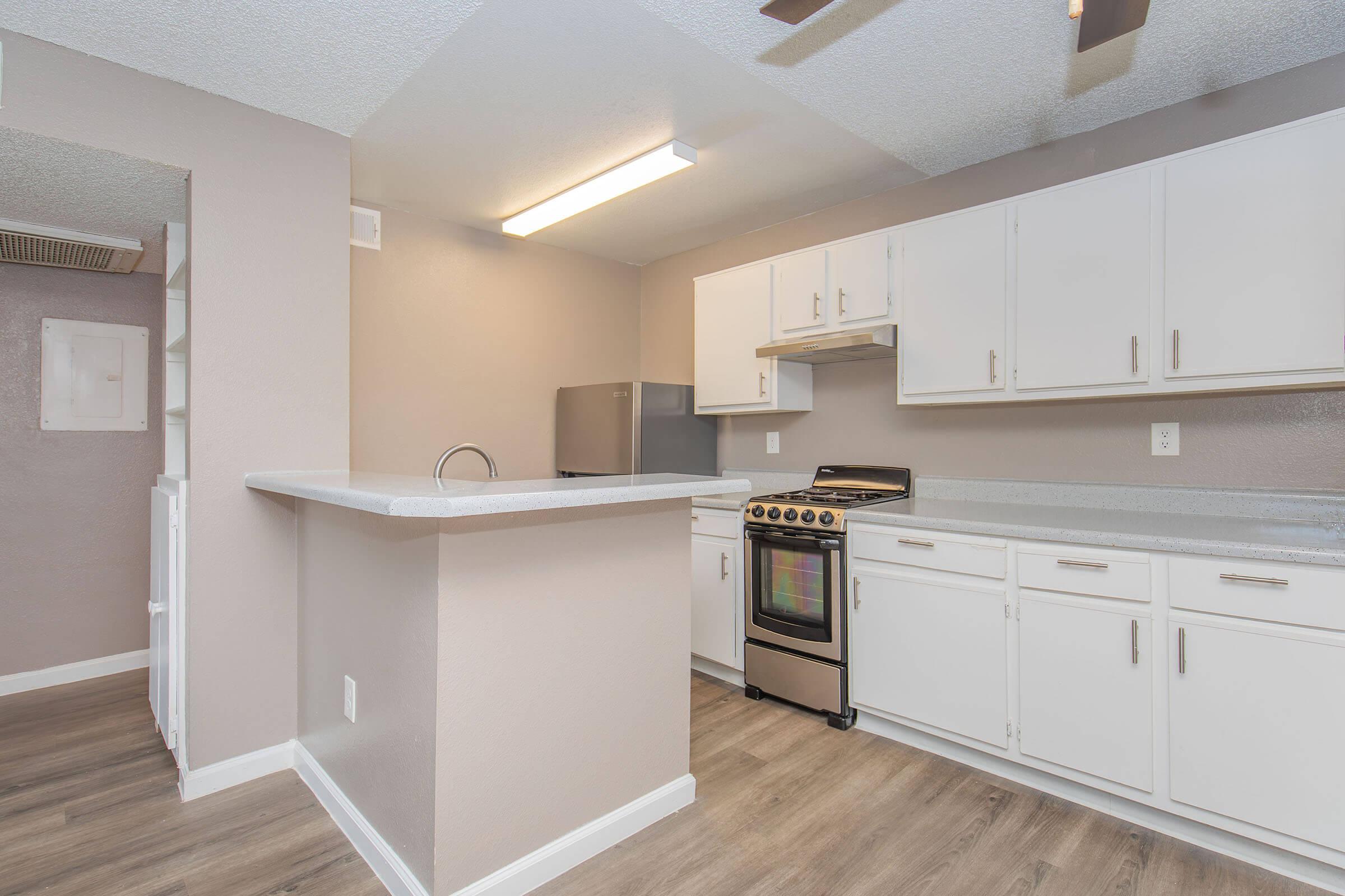 Modern kitchen with white cabinetry, stainless steel stove, and a refrigerator. The countertops are light gray, and the flooring is a wood-like laminate. A ceiling fan adds to the contemporary feel, and there's an open layout with a low bar countertop separating the kitchen from the living area.