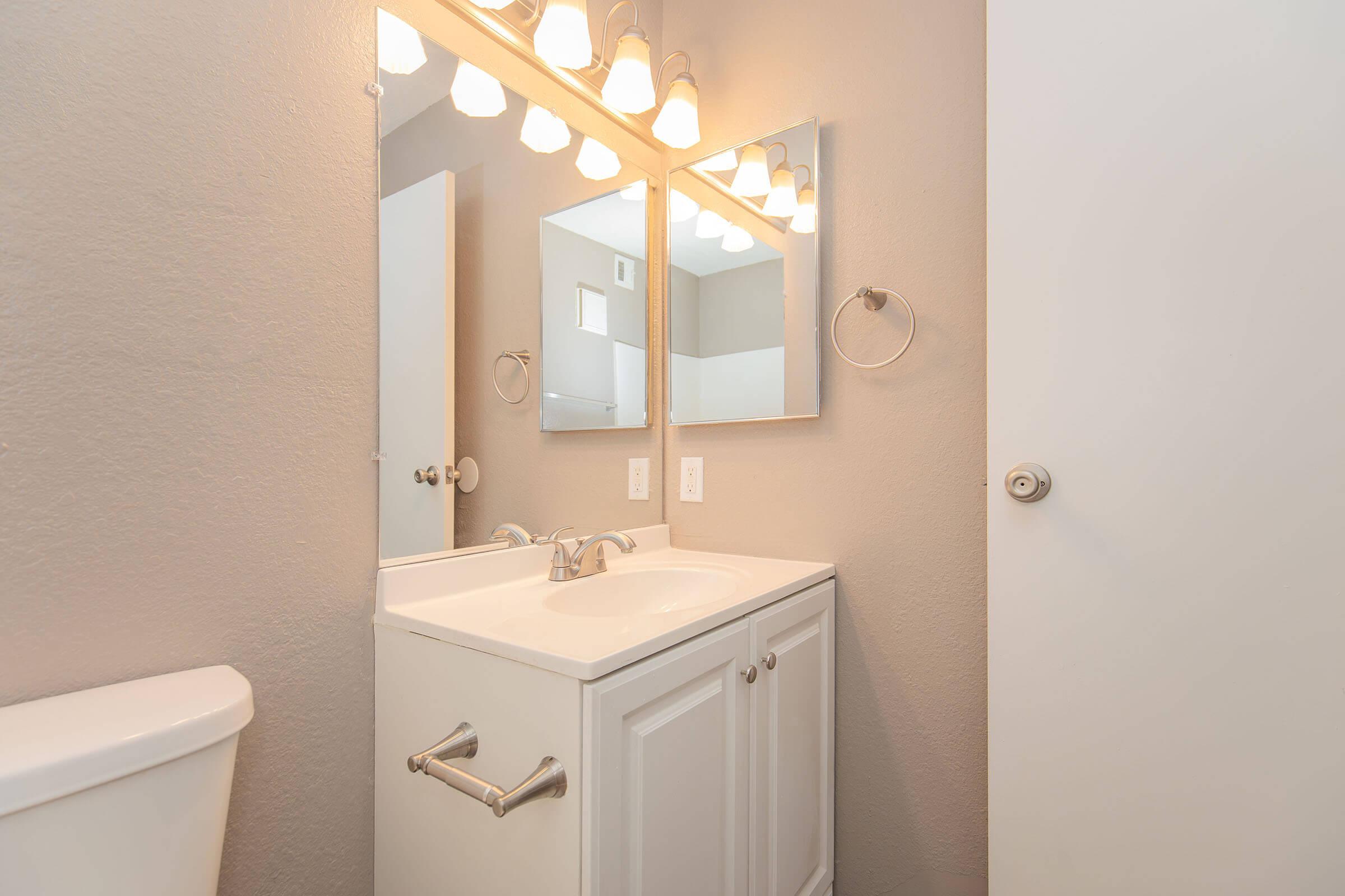 A well-lit bathroom featuring a white sink cabinet with a countertop, two mirrors above the sink, and modern light fixtures. The walls are painted a light gray, and a white toilet is visible on the left. There is a towel ring next to the sink and a door partially open in the background.