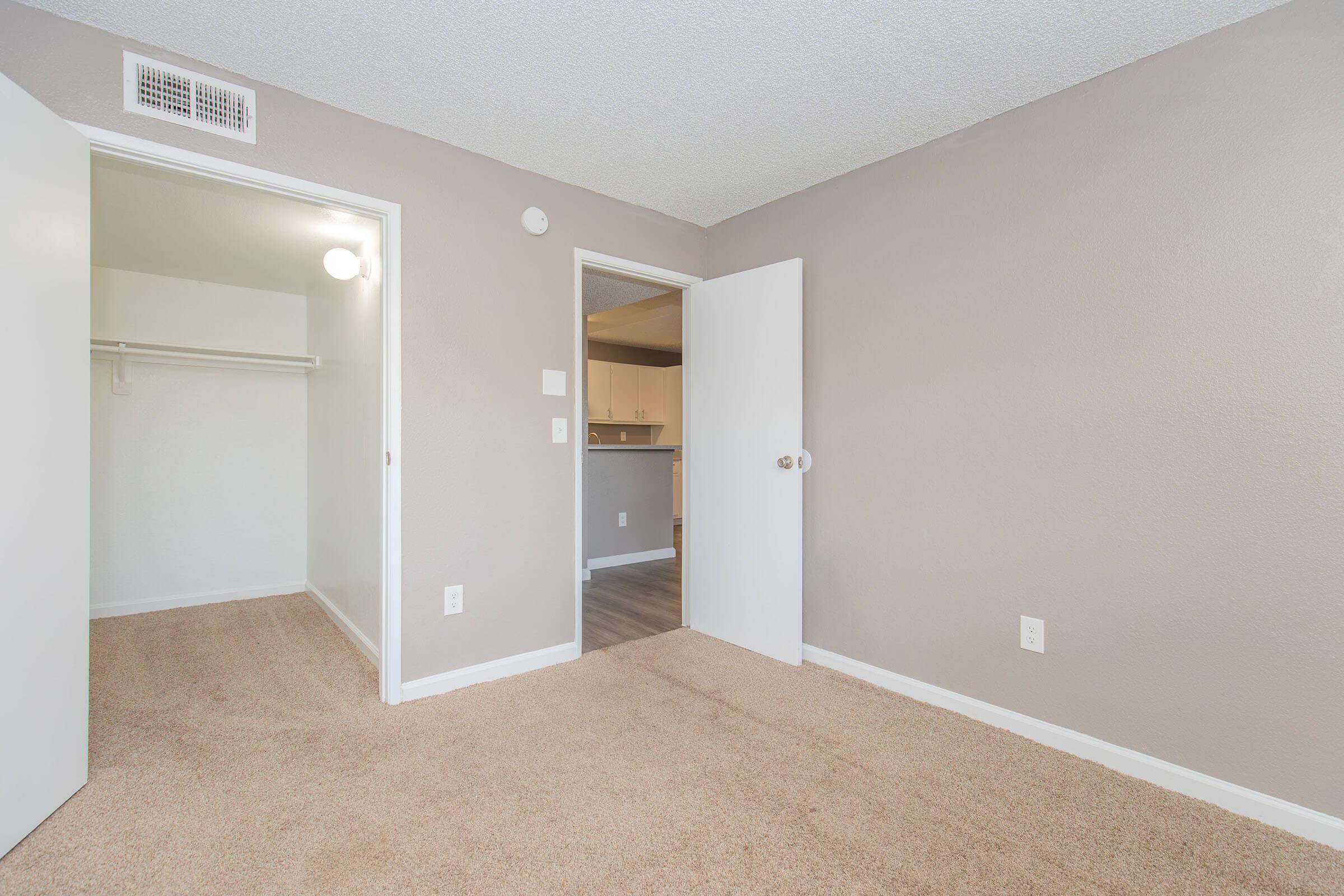 A spacious, empty bedroom with light beige walls and carpet. There are two doors: one leading to a closet and the other to another room. A light fixture hangs from the ceiling, providing a bright atmosphere. The overall space is clean and ready for furnishing.