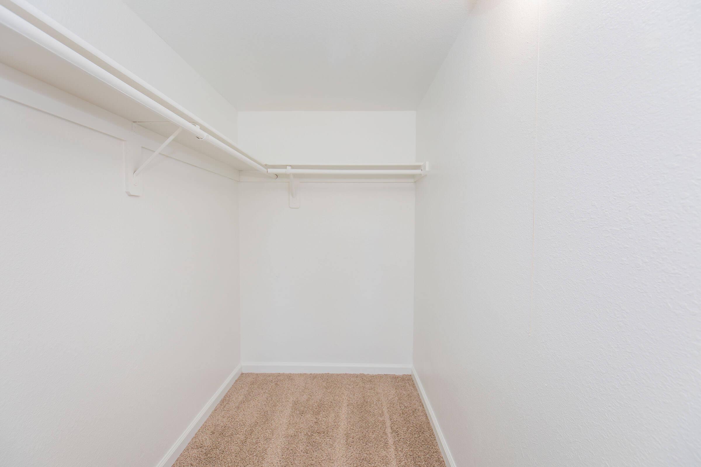 A vacant walk-in closet with white walls and a light brown carpet. It features two horizontal closet rods for hanging clothes, providing ample space for storage. The overall atmosphere is clean and minimalistic, emphasizing the emptiness of the space.
