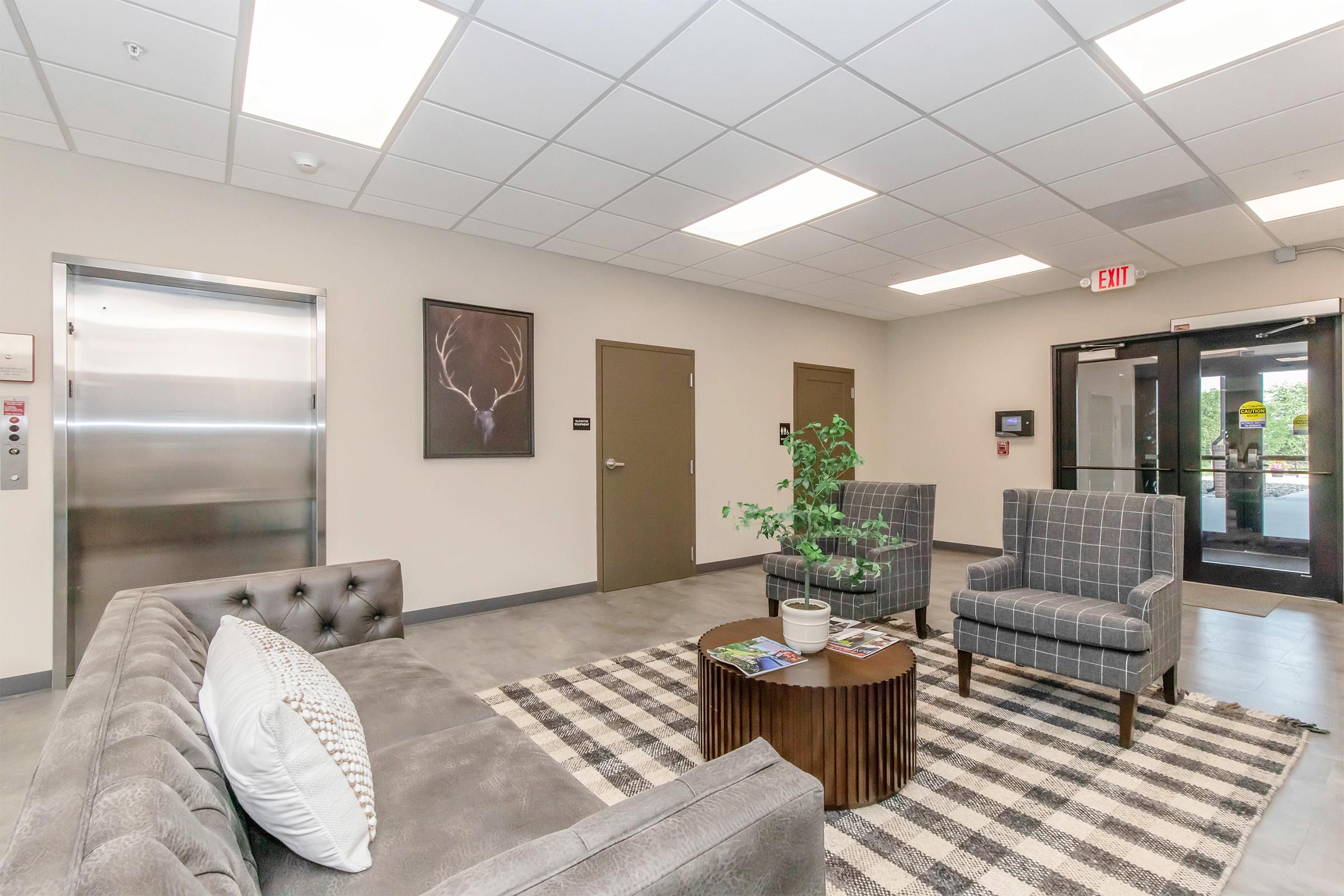 A modern lobby area featuring a gray sofa with decorative pillows, two checkered armchairs, and a small table with magazines. There's a potted plant on the table, a wall art piece of a deer antler, an elevator on the left, and glass doors leading outside. The flooring is gray and the overall design is minimalist and inviting.