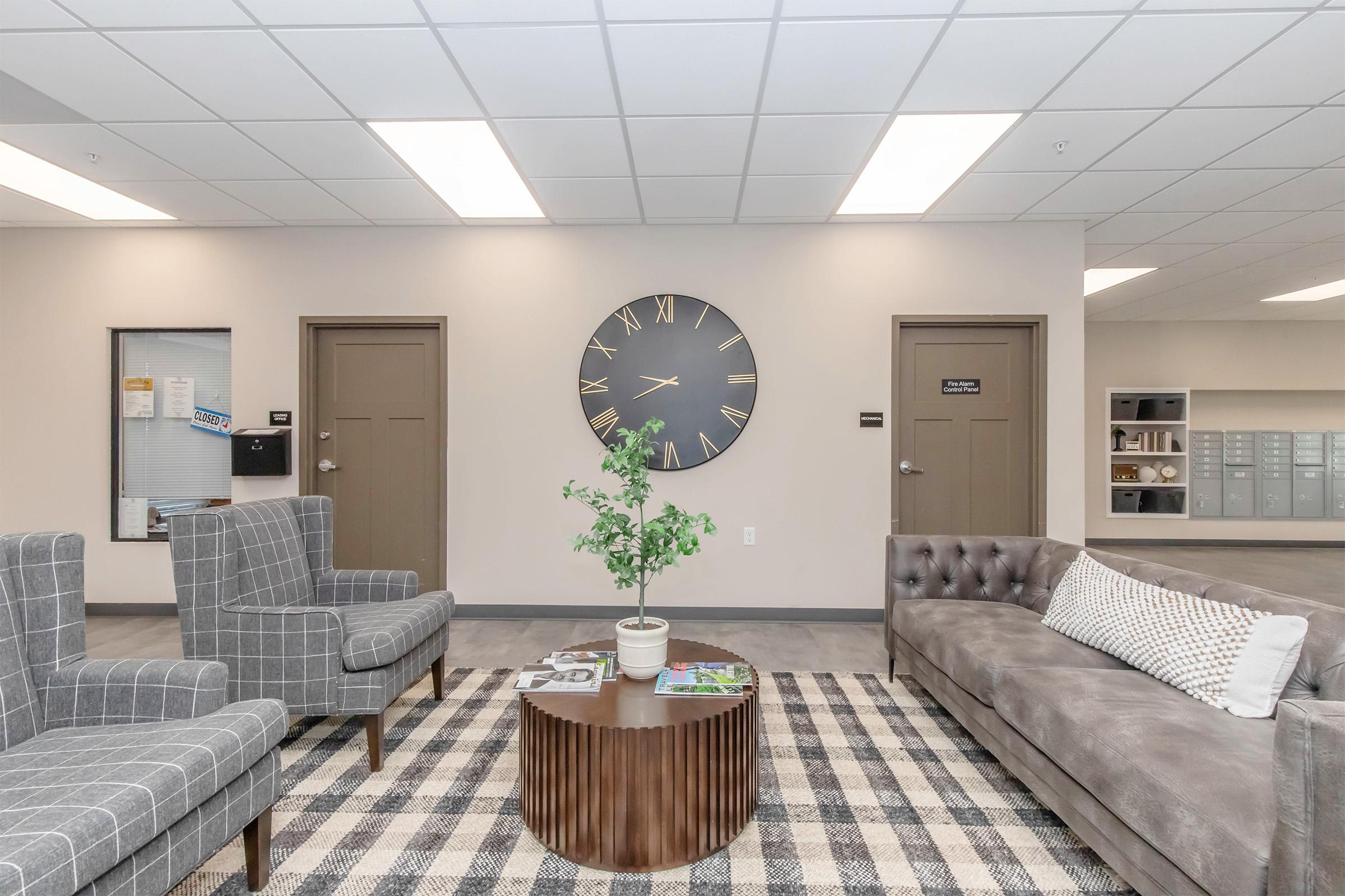 A modern waiting area featuring two patterned armchairs and a plush sofa with a decorative pillow. In the center, a round table supports a small potted plant. A large wall clock and two doors are visible, along with a bulletin board and storage units in the background. The space has a neutral color palette and bright lighting.