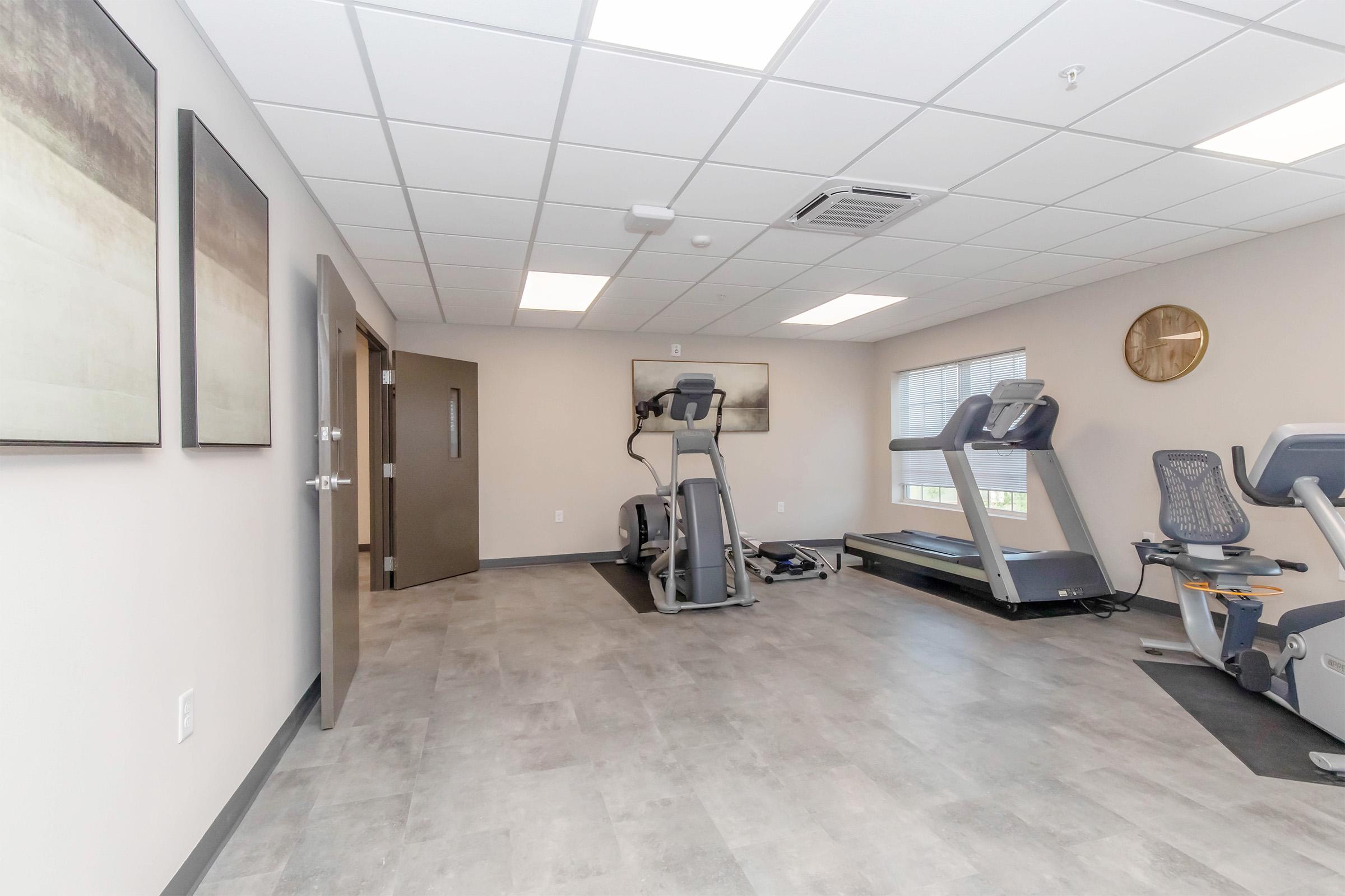 A spacious fitness room featuring two exercise machines, a treadmill, and an elliptical trainer. The room has neutral-colored walls, modern artwork, and large windows allowing for natural light. There are two doors, one leading outside and another to an adjacent area.