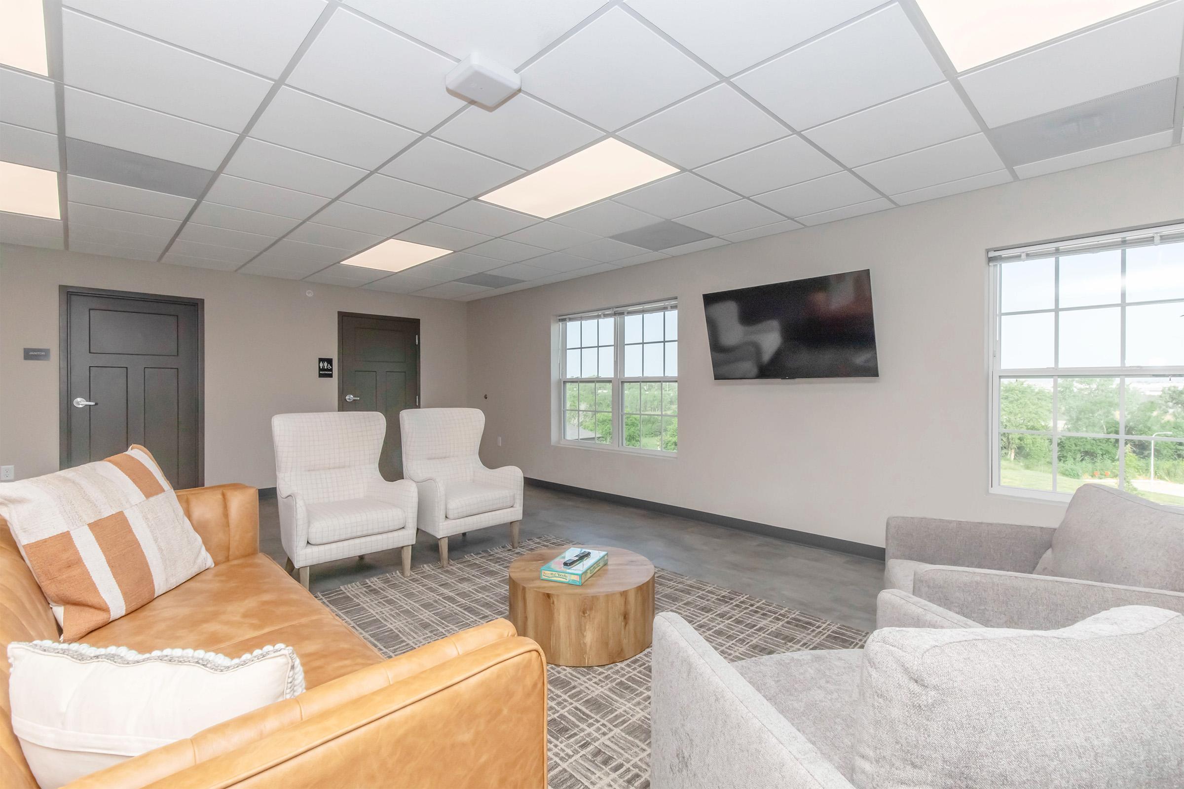 A modern lounge area featuring two light-colored armchairs, a brown leather sofa, a round wooden coffee table, and a gray area rug. Large windows allow natural light, and a flat-screen TV is mounted on the wall. The room has a neutral color scheme with a simple, contemporary design.