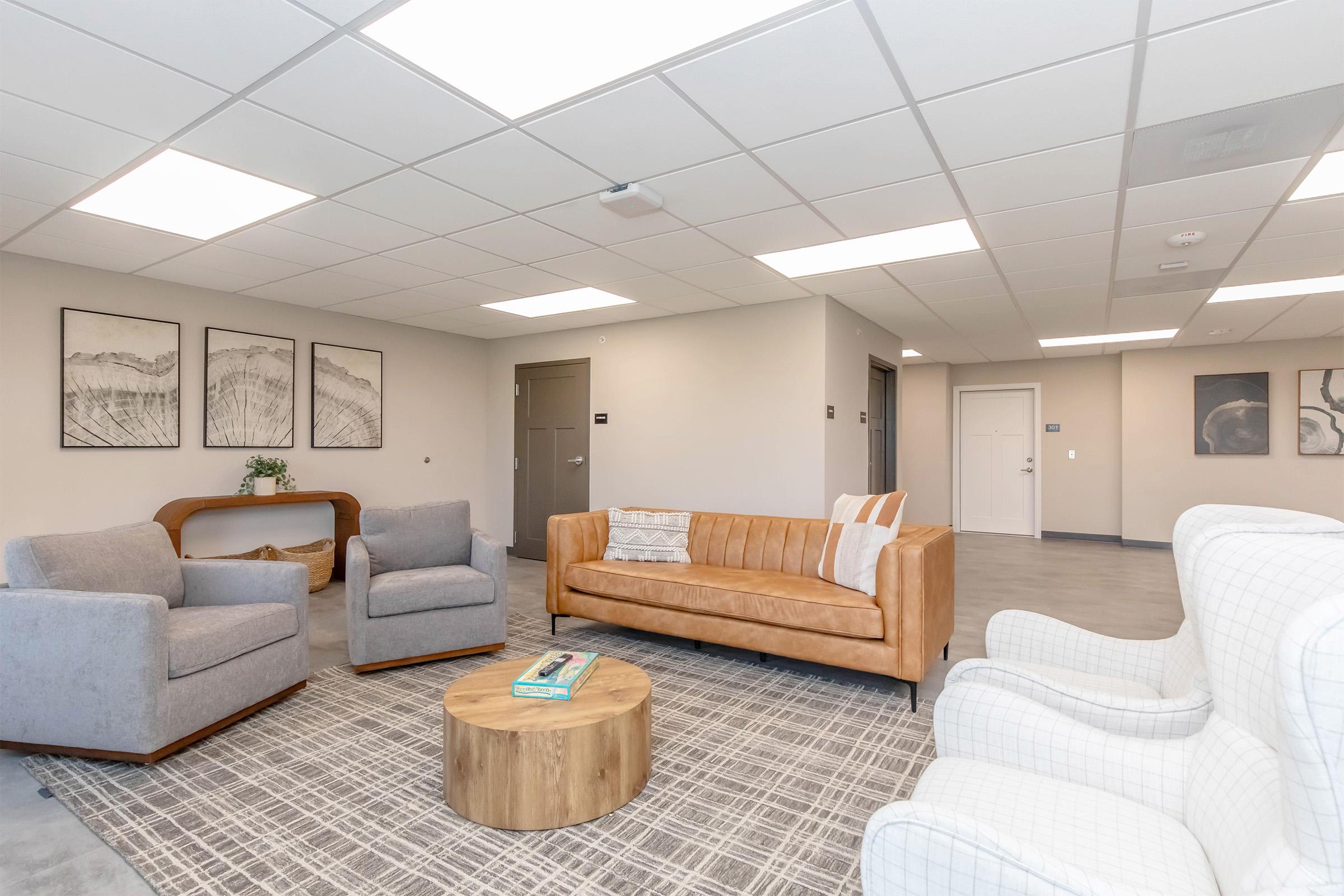 A modern, well-lit lounge area featuring two gray upholstered armchairs, a stylish tan leather sofa, a round wooden coffee table, and a patterned area rug. The walls are adorned with framed artwork, and there are doors leading to other rooms in the background.