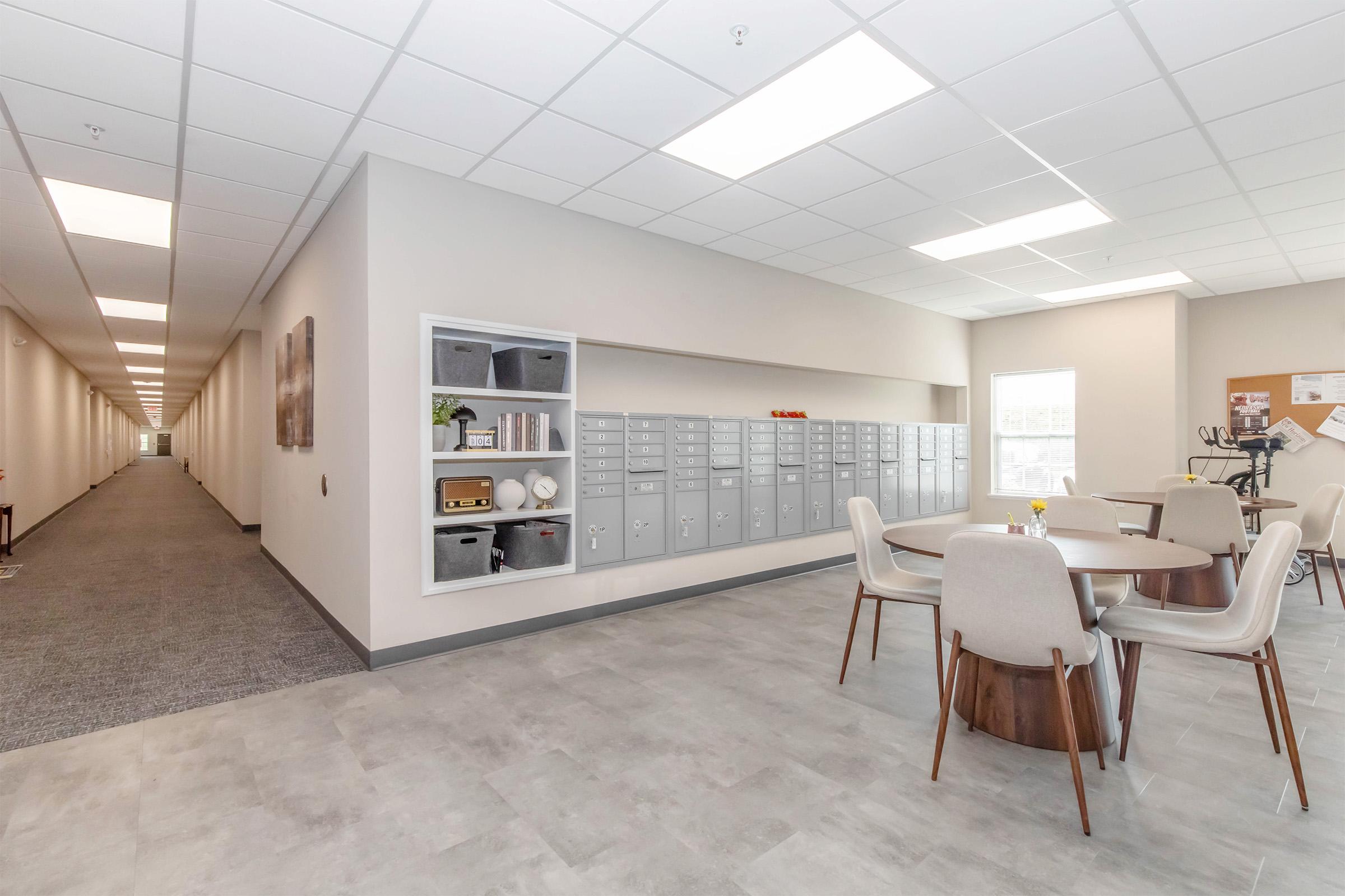 A modern interior of a hallway with mailboxes on one wall. There is a round dining table with four chairs in the foreground, and an organized wall with a bulletin board and some decorative elements. The space is well-lit, featuring neutral colors and a clean, open design.