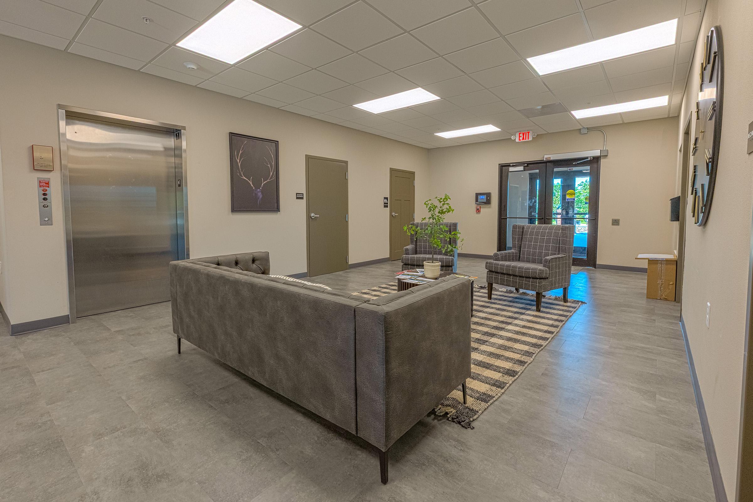 Modern lobby area featuring a gray couch and two patterned armchairs on a striped rug. There are elevator doors on one side and two office doors on the opposite wall. Natural light enters through large glass doors leading outside, and there are minimal decorations, including framed art.