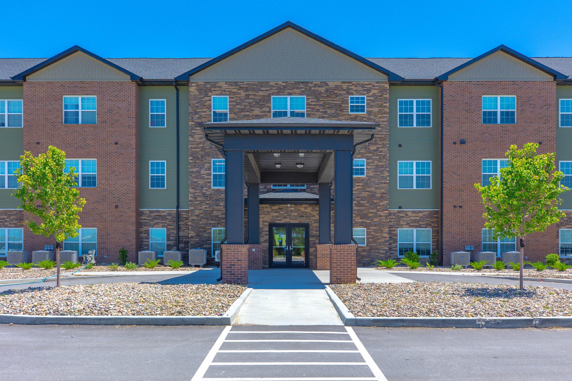 A modern multi-story apartment building with a brick and green exterior, featuring a covered entrance. The entrance is framed by two small trees, and there is a well-maintained landscape with gravel and shrubs. The sky is clear and blue, enhancing the appeal of the property.