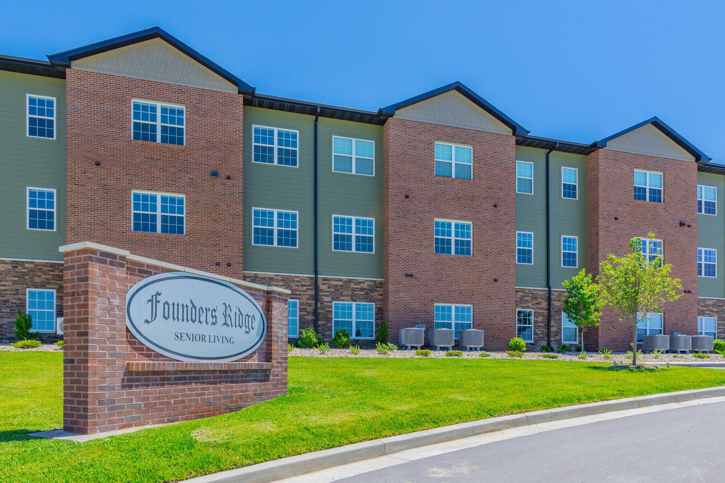 Exterior view of Founders Ridge Senior Living, a modern multi-story brick building with green siding. The sign prominently displays the name of the facility, and the landscaped area features grass and small trees. The sky is clear and blue, creating a bright and inviting atmosphere.
