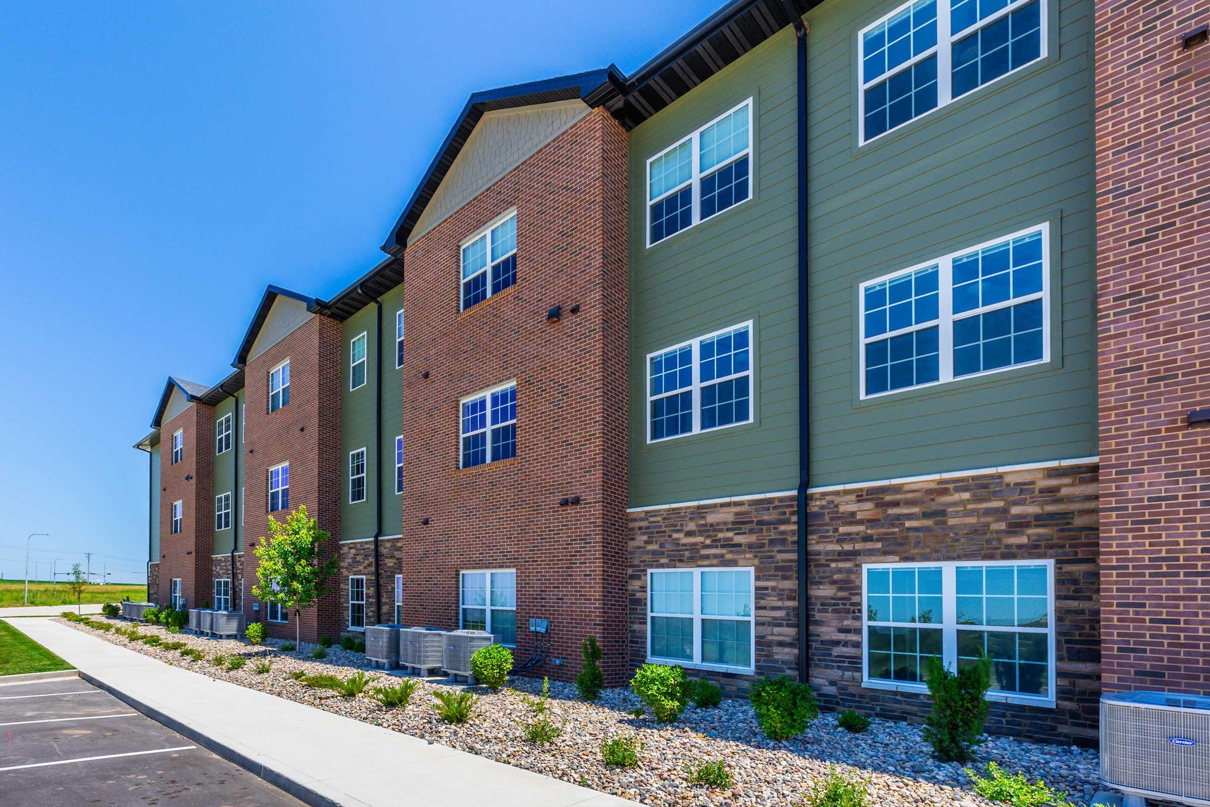 A modern apartment building with a mix of brick and green siding. The structure features large windows and well-maintained landscaping, including small shrubs and rocks along the walkway. The sky is clear and blue, highlighting the vibrant colors of the exterior.
