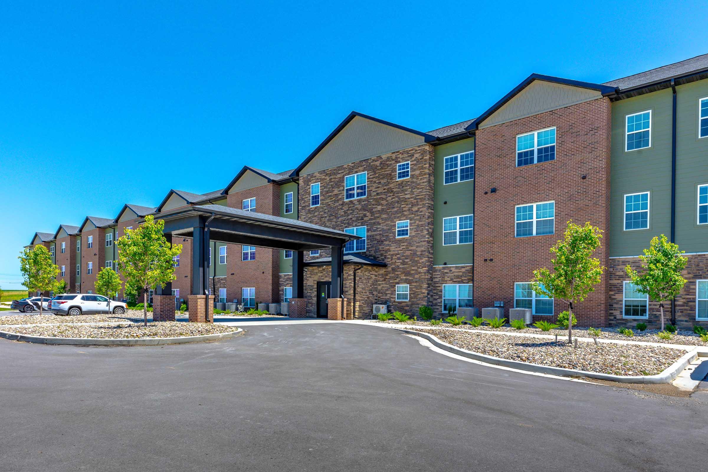 A modern multi-story building with a brick and siding exterior, featuring a covered entrance. The landscape includes small, manicured trees and flower beds. A paved parking area is visible in front, with a few parked cars and a clear blue sky overhead.
