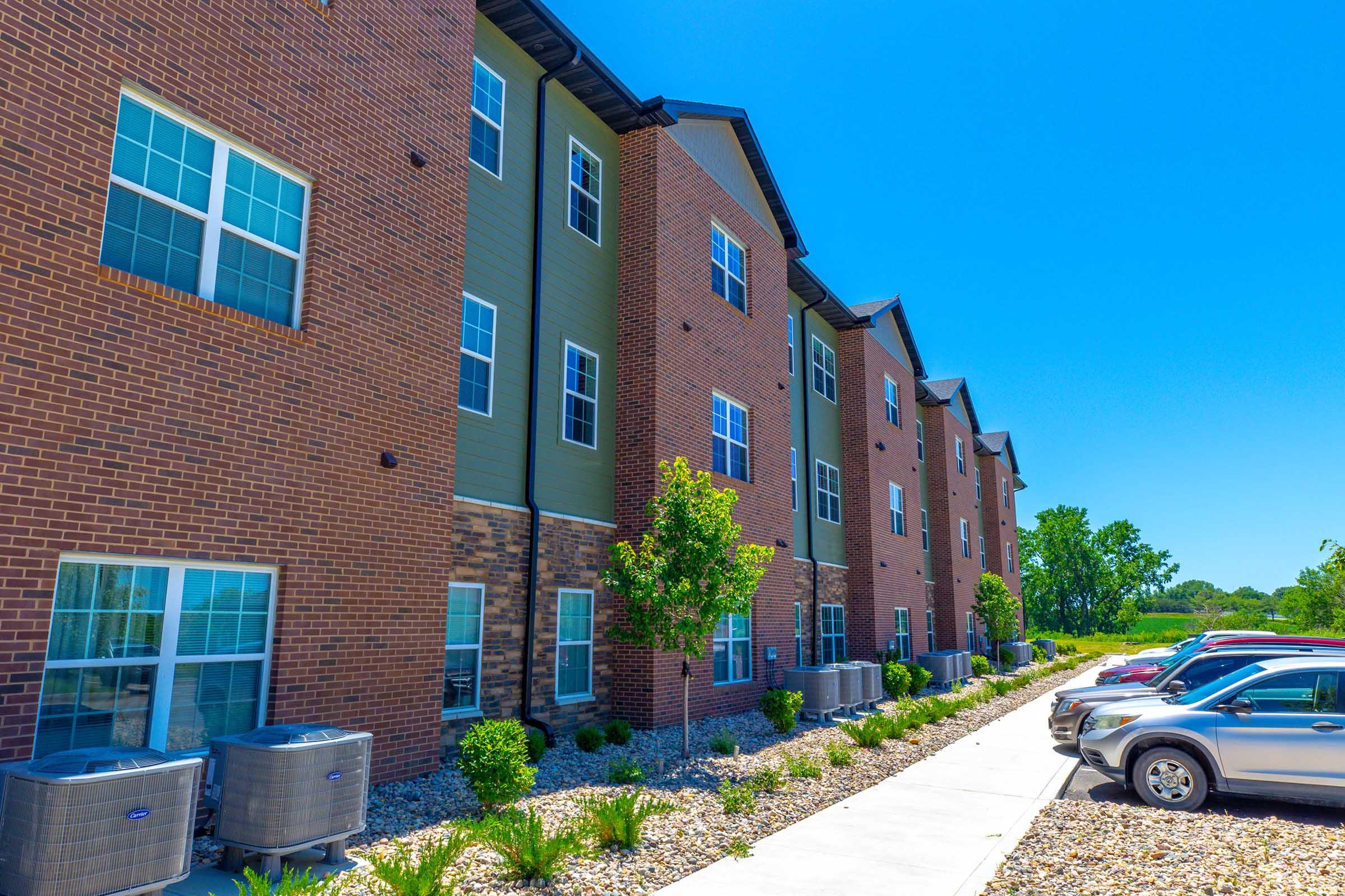 A modern multi-story brick and siding apartment building with well-maintained landscaping, including shrubs and small trees. Air conditioning units are visible along the side of the building, and a paved walkway leads to parked cars on the right under a clear blue sky.