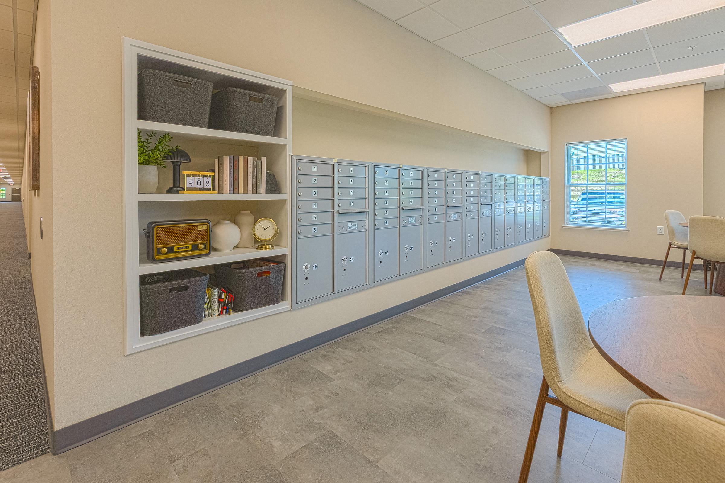 A modern interior space featuring a row of silver mailboxes mounted on the wall. To the left, there's a white shelf with decorative items, including storage baskets, books, and a vintage-style radio. The flooring is a light gray tile, and large windows allow natural light to fill the room.