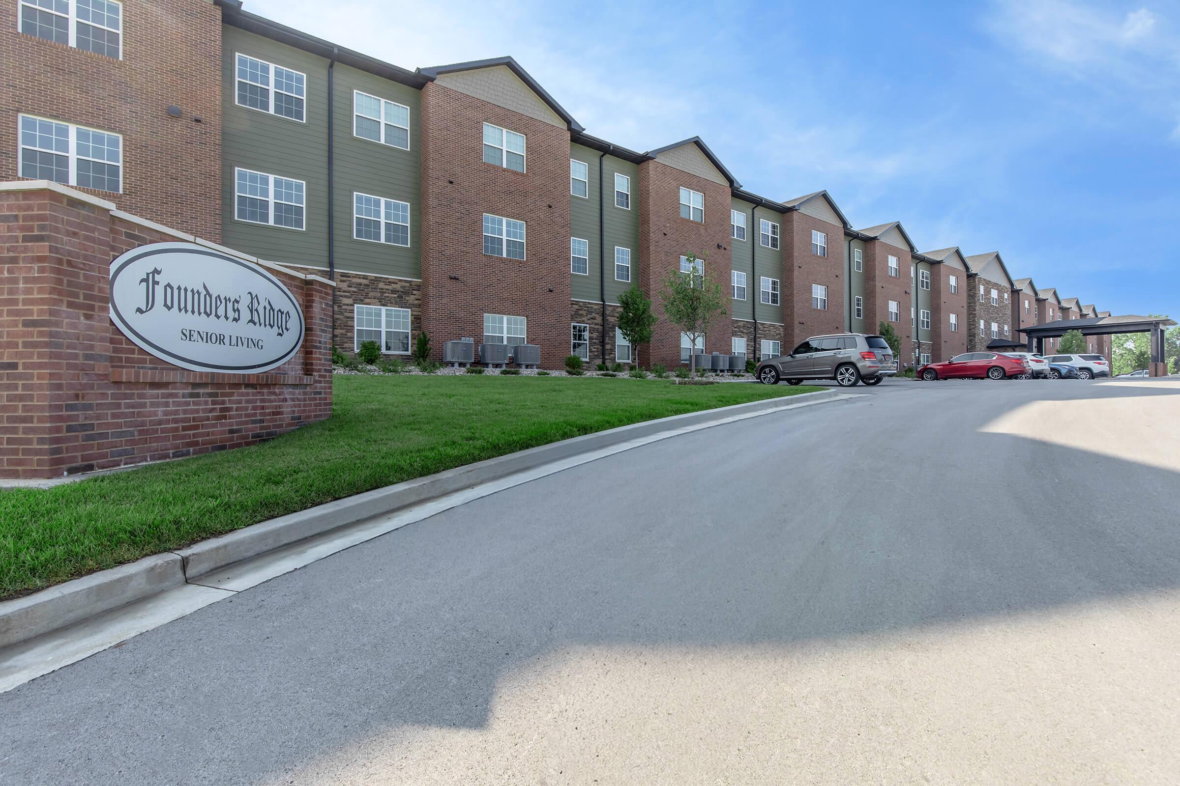 Exterior view of Founder's Ridge Senior Living facility, showcasing a modern brick building with multiple windows, landscaped green areas, and a paved driveway. Vehicles are parked along the side, and a sign with the name of the facility is prominently displayed at the entrance. Blue sky in the background.