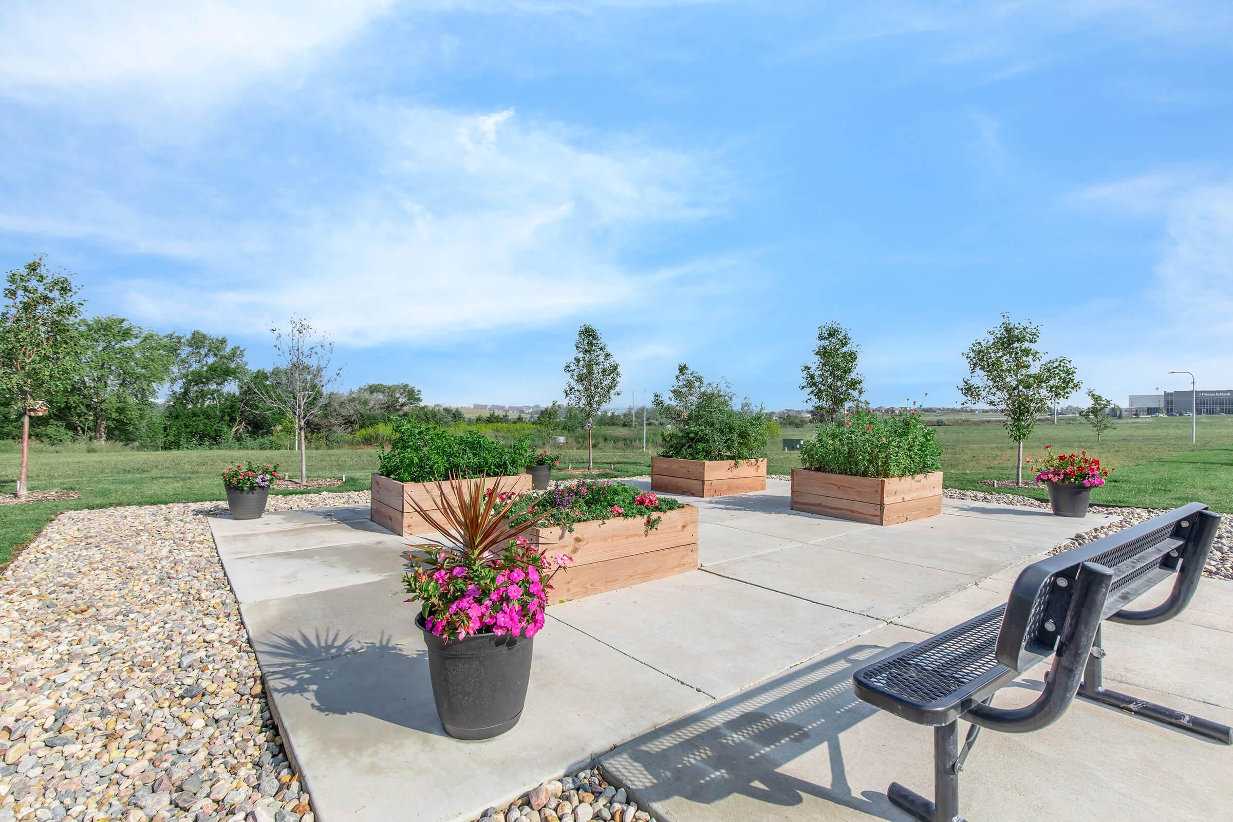 A tranquil garden area featuring raised wooden planter boxes filled with vibrant flowers and greenery. Surrounding the planters are flower pots and a paved seating area with a bench, set against a clear blue sky and open grassy landscape. Trees are visible in the background.