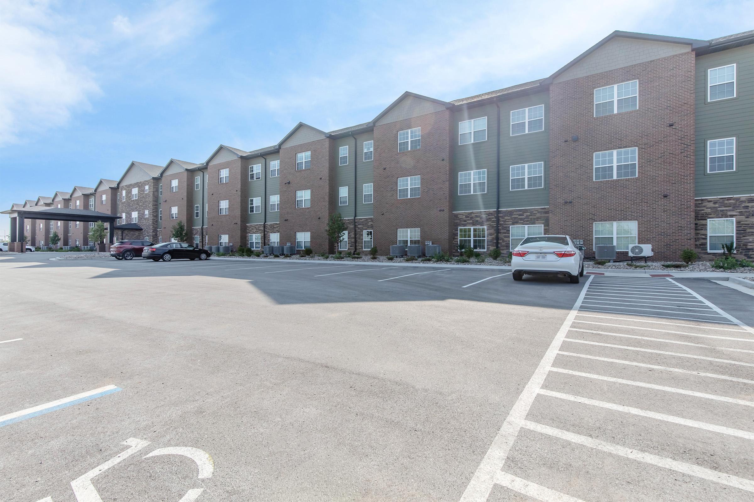 Modern brick apartment building with multiple floors and a flat roof. The facade features a mix of dark brick and green siding. A well-maintained parking lot is in front, with several vehicles parked. The sky is clear and blue, enhancing the inviting atmosphere of the property.