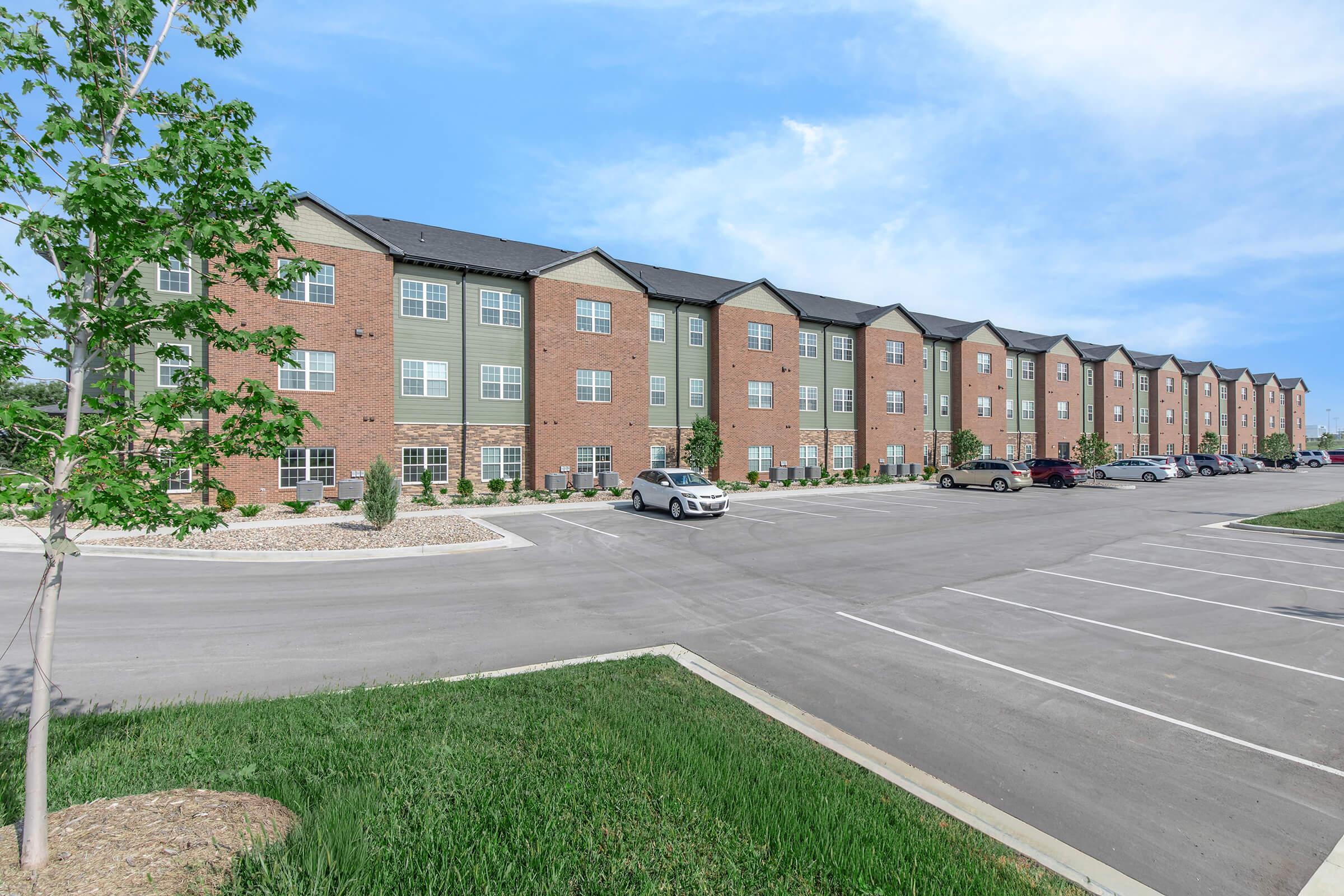 A row of modern multi-story apartment buildings with brick and siding exteriors. The scene features a neatly paved parking lot with several cars parked. Lush green grass and small trees line the foreground, with a clear blue sky above.
