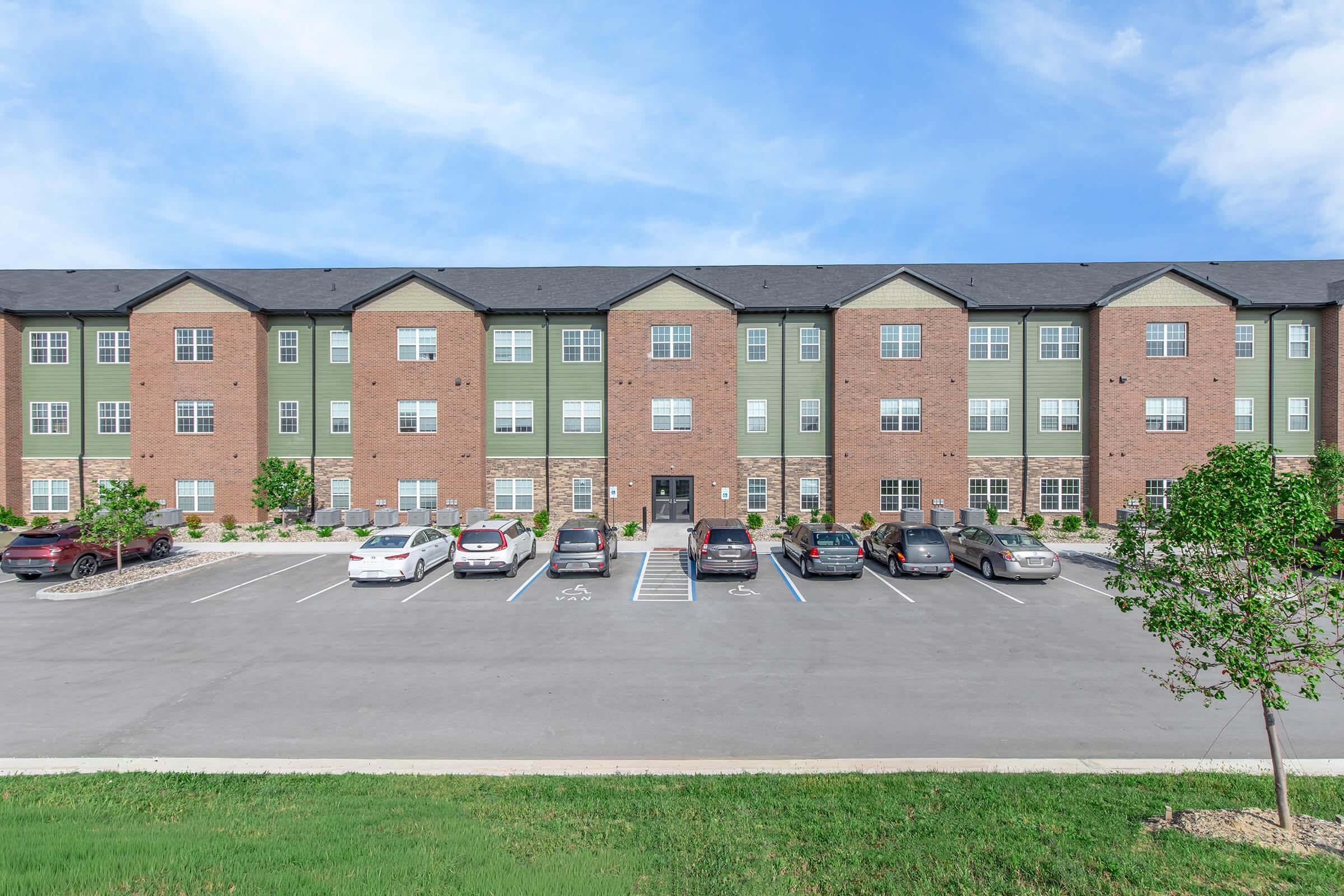 A modern three-story apartment building with a mix of brick and green siding located in a well-maintained area. Several cars are parked in designated spaces in the foreground, and there are trees and grassy areas surrounding the building under a clear blue sky.