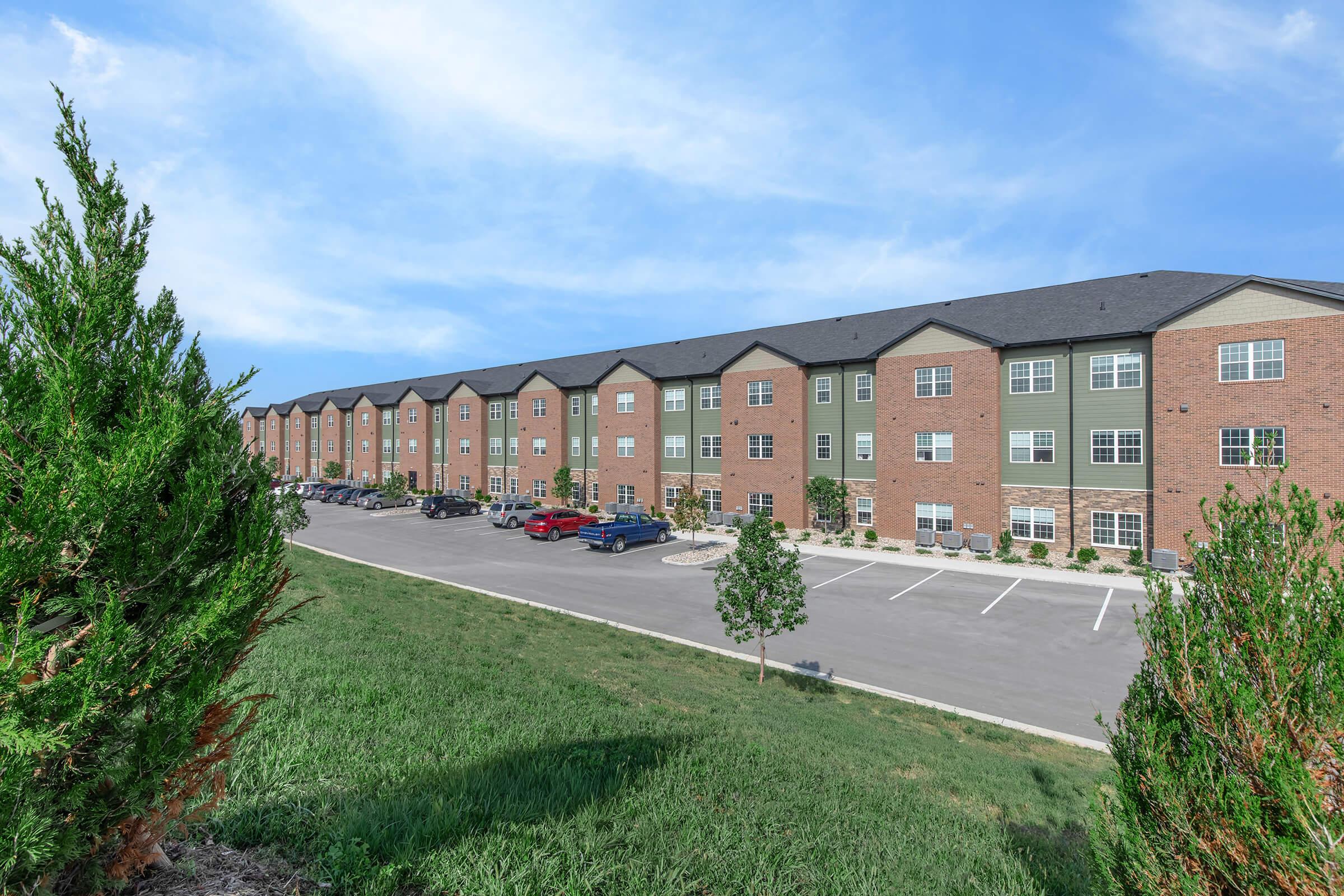 A row of modern apartment buildings featuring brick and green exteriors, with a well-maintained parking lot in front. Lush green grass is visible in the foreground, and the sky is bright with a few clouds.