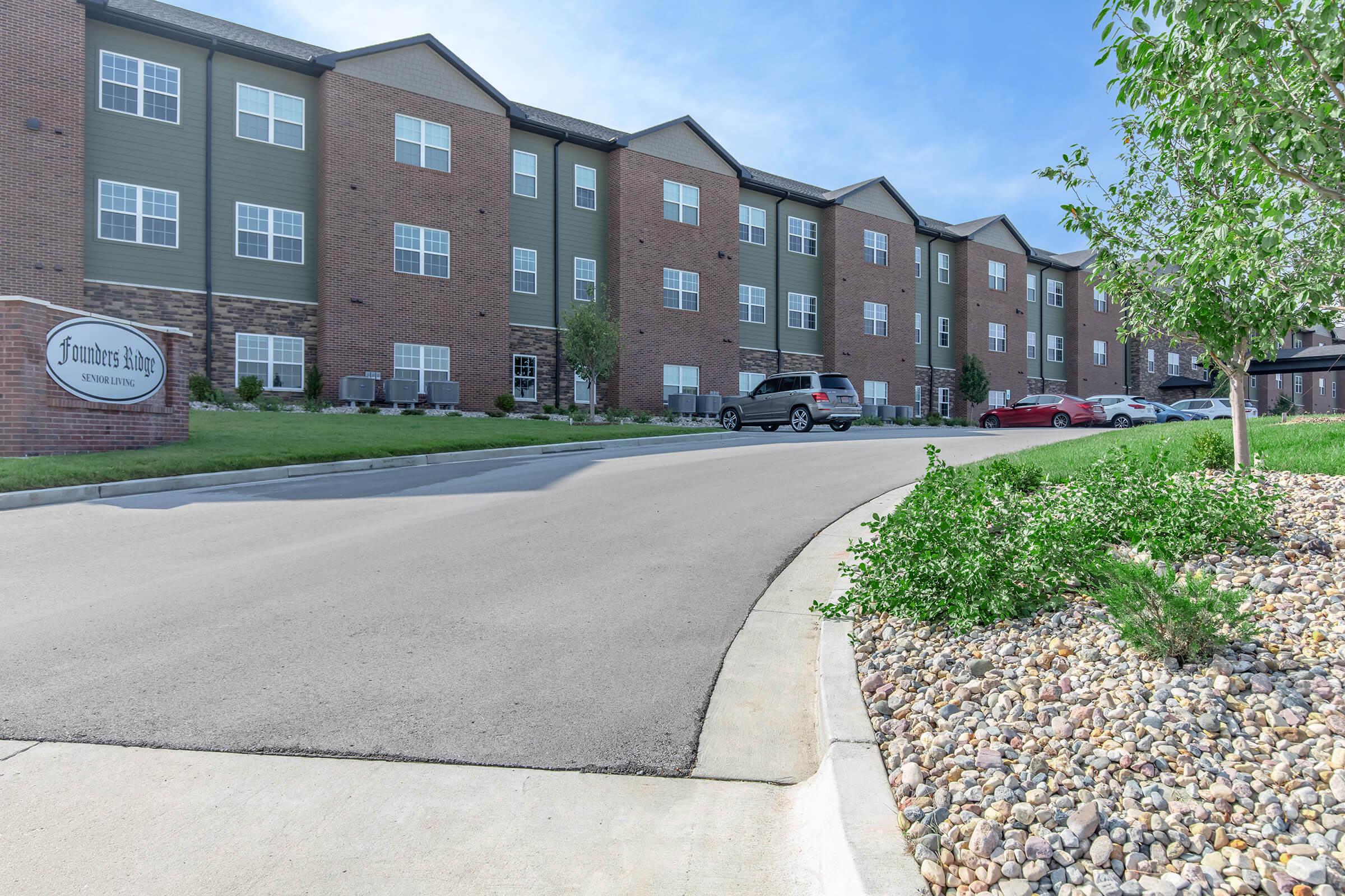 Street view of a multi-story apartment building with a brick exterior. A sign reads "Jasmine's Place." The parking lot features several vehicles and landscaped areas with greenery and gravel. Clear blue sky in the background.