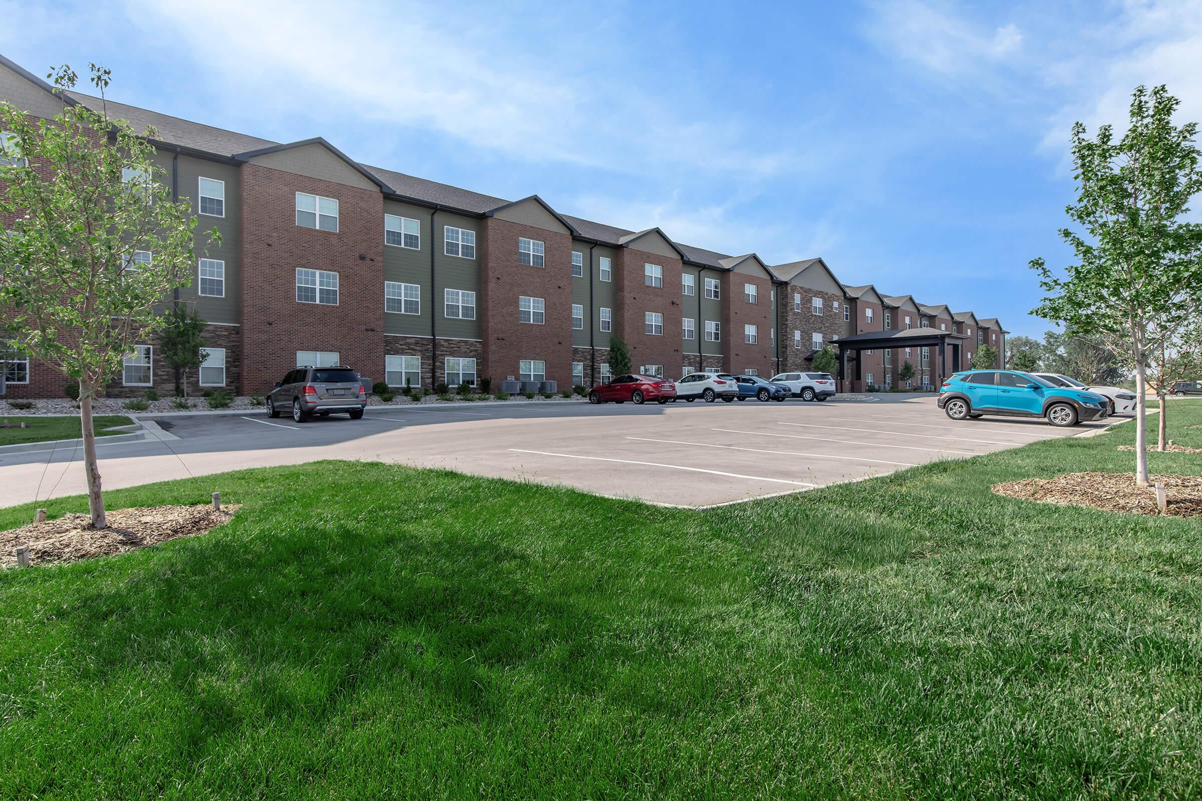 A modern multi-story brick apartment building with multiple windows and parked cars in the foreground. The surrounding area features well-maintained green grass and small trees. The sky is clear with a few clouds.