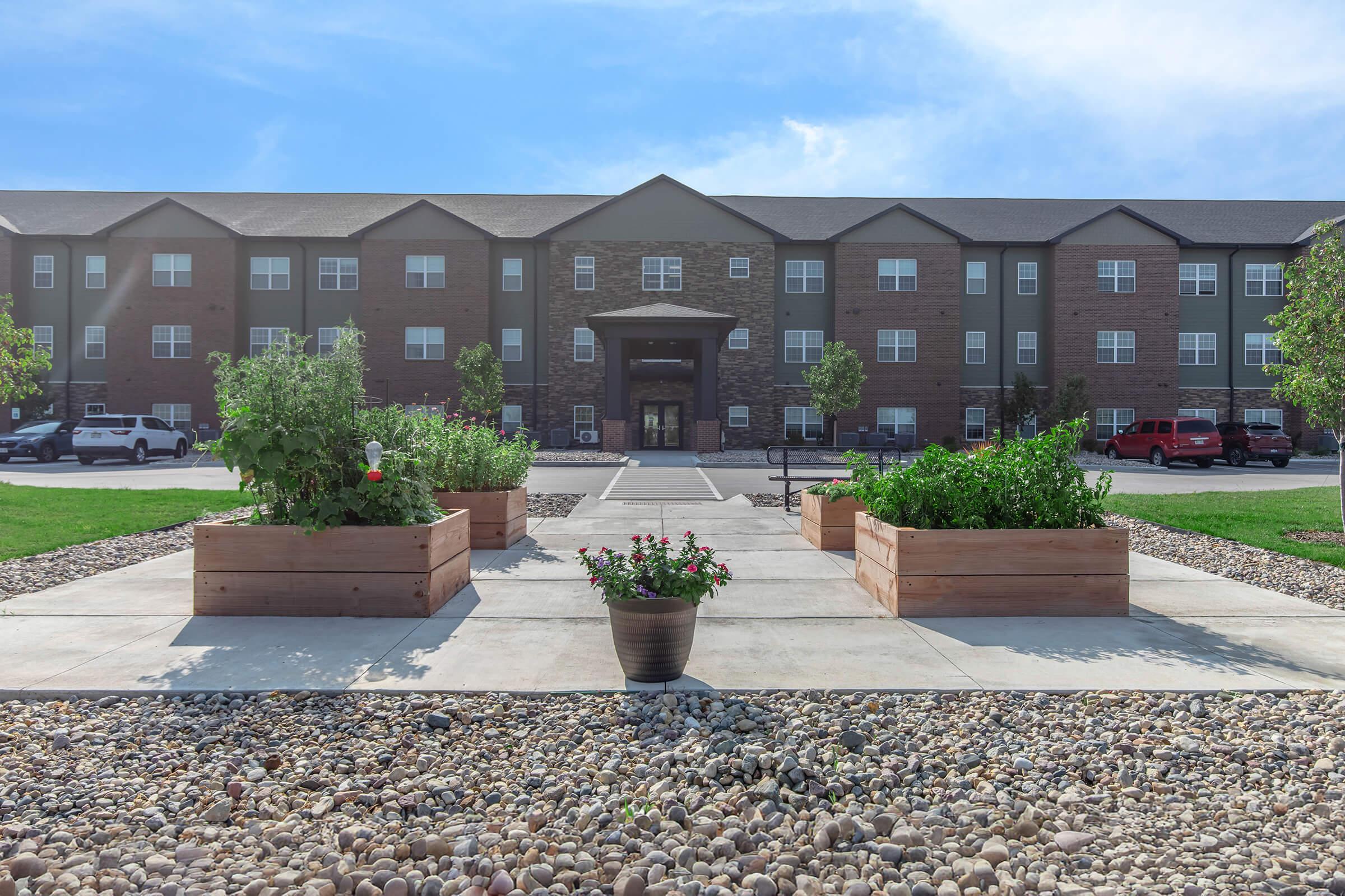 Exterior view of a modern apartment building with a landscaped entrance. In the foreground, there are raised garden beds with green plants and flowers, surrounded by decorative gravel. The building features a mix of brick and siding, with a clear blue sky above. Parking spaces are visible in front of the building.