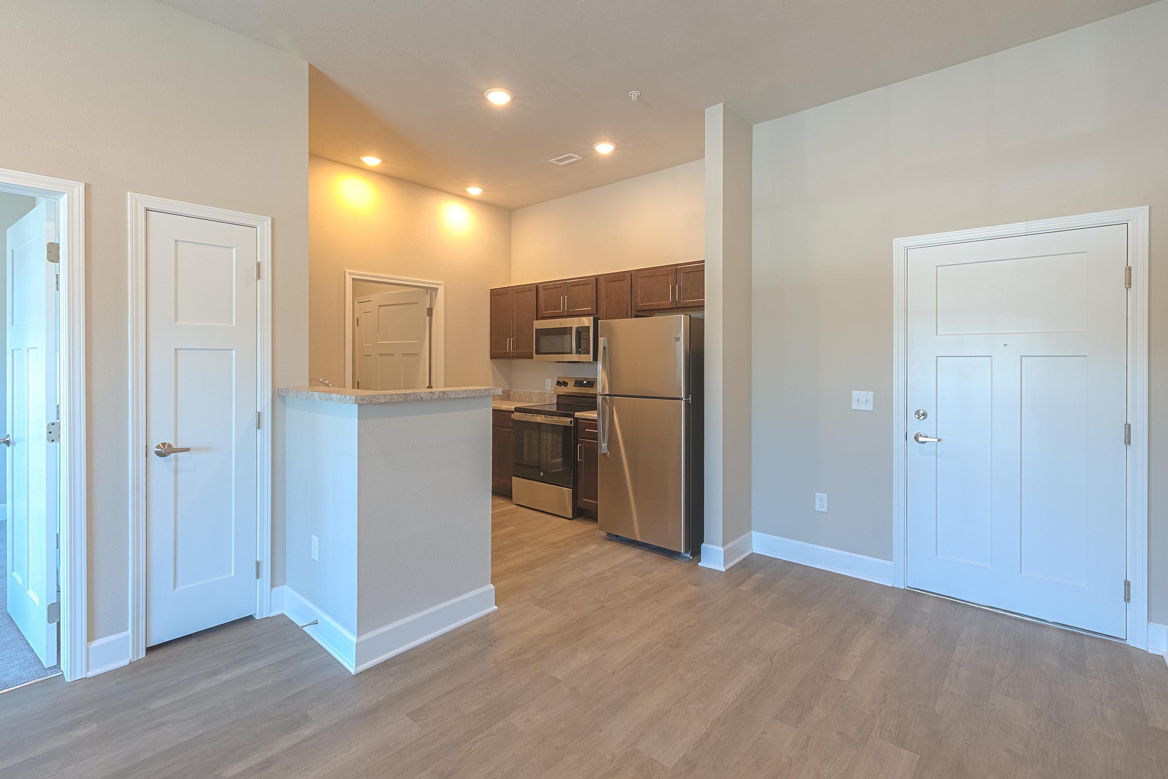 A modern apartment interior showcasing an open living space with light-colored walls, wooden laminate floors, and a kitchen area featuring stainless steel appliances, including a refrigerator and oven. The layout includes a doorway leading to an entrance, with abundant natural light illuminating the room.