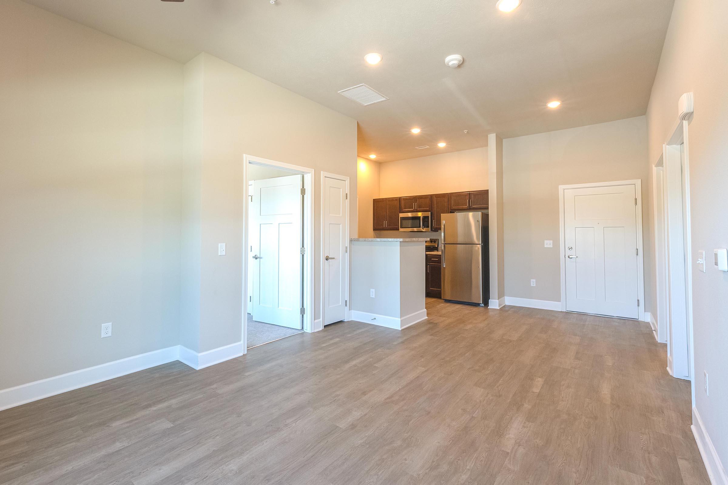 A bright and spacious living area with light-colored walls and wood-like flooring. The room features an open layout leading to a contemporary kitchen with stainless steel appliances. There are two doorways, one leading to a closet or bathroom, and another to the outside. Natural light streams in from the windows.