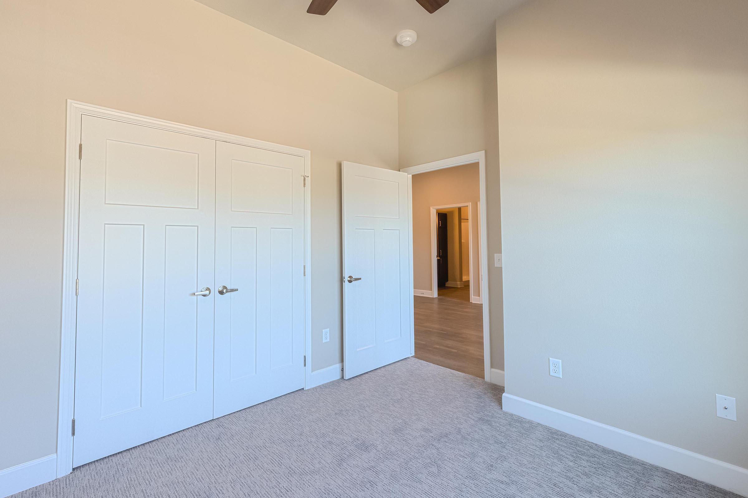 A bright, empty room with light beige walls and a carpeted floor. There are two closed white doors leading to closets and one open door in the background, suggesting access to another room. A ceiling fan is visible, and the space has a clean, modern aesthetic.