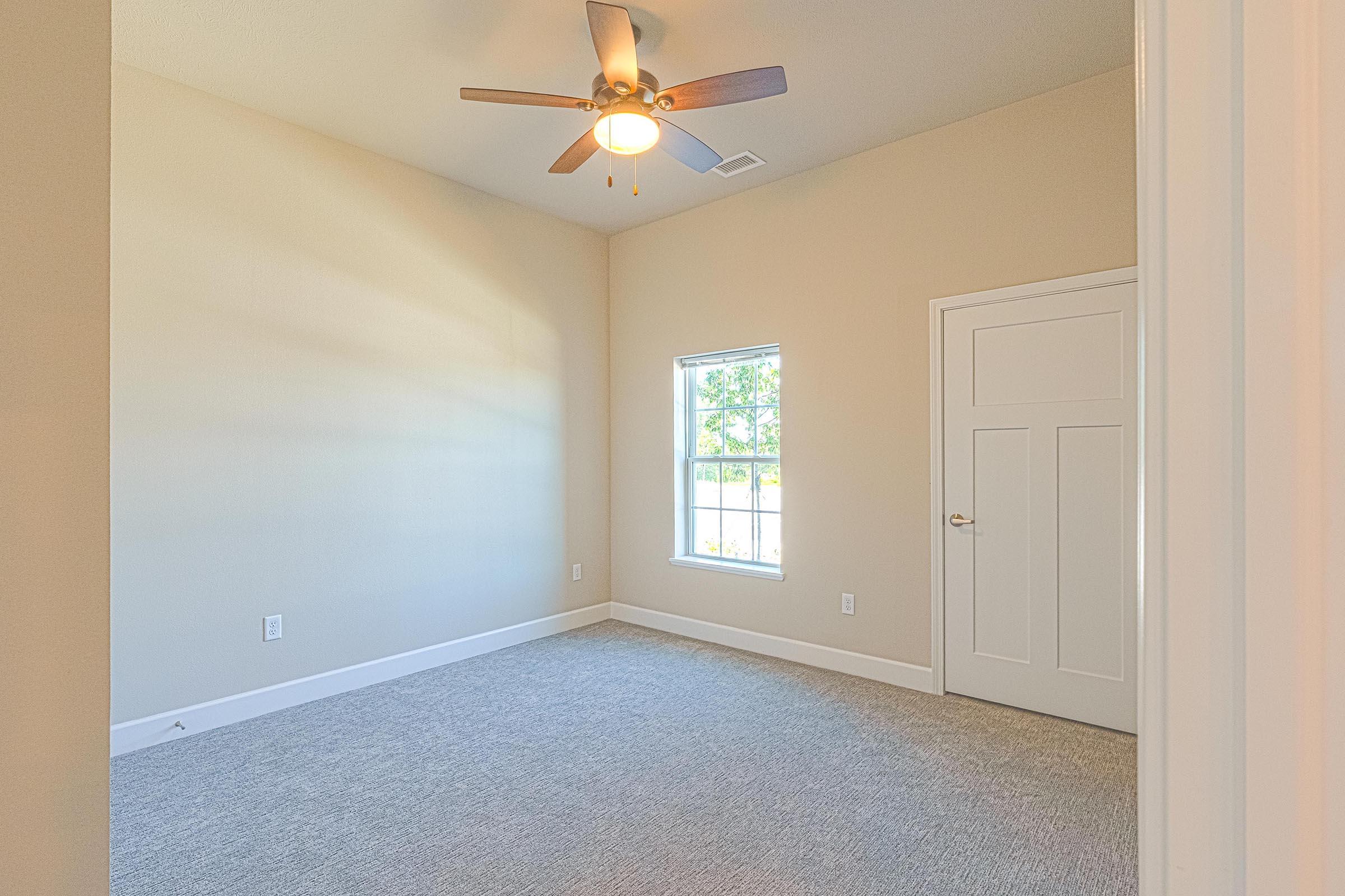 A well-lit, empty room featuring light beige walls and carpeted flooring. There is a ceiling fan with wooden blades, a window allowing natural light in, and a closet door. The overall ambiance is spacious and clean, suitable for various uses such as a bedroom or office.